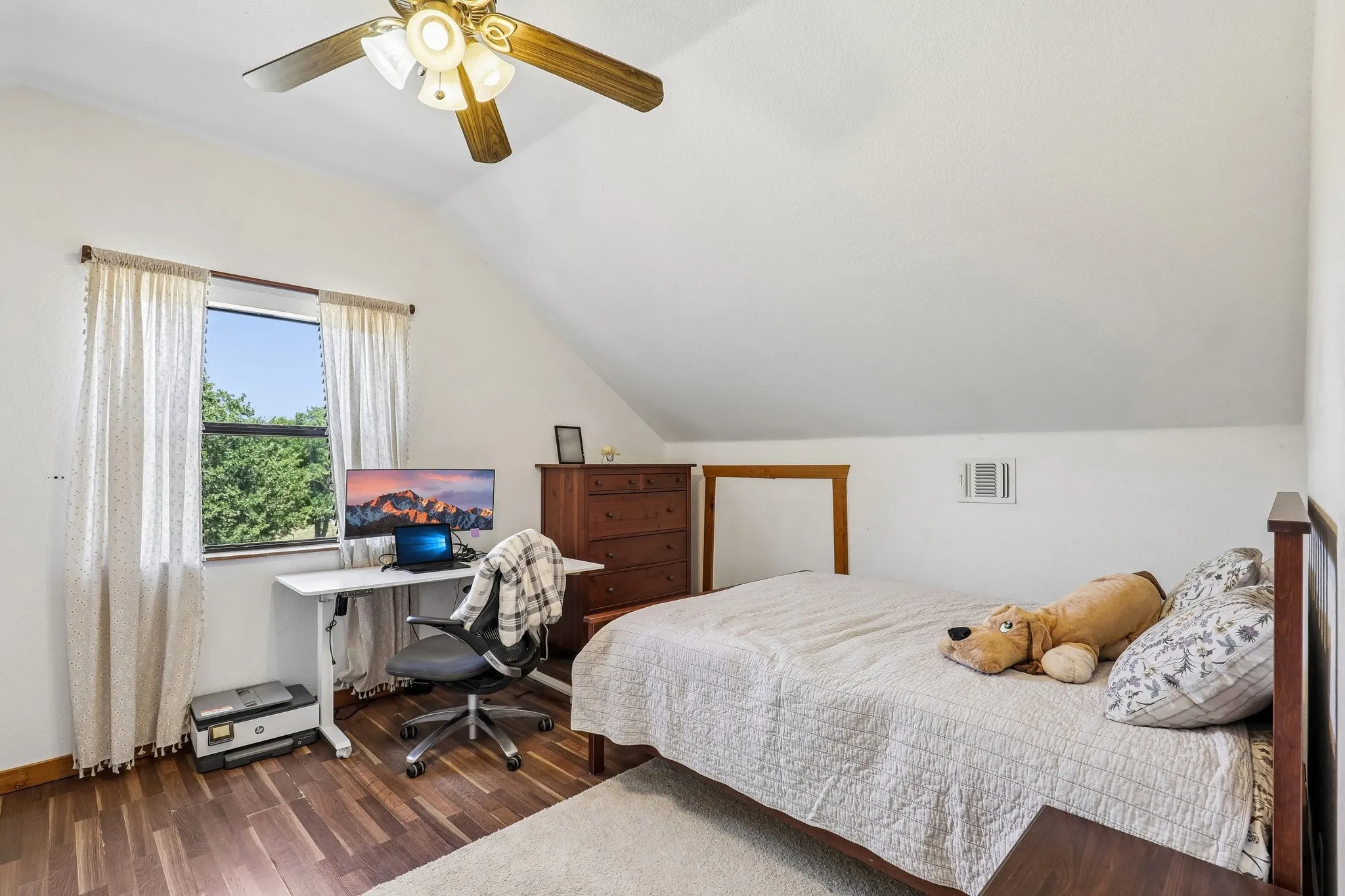 Room featuring an angled ceiling, wood-finish flooring, a window with exterior tree views, and a ceiling fan with integrated lighting