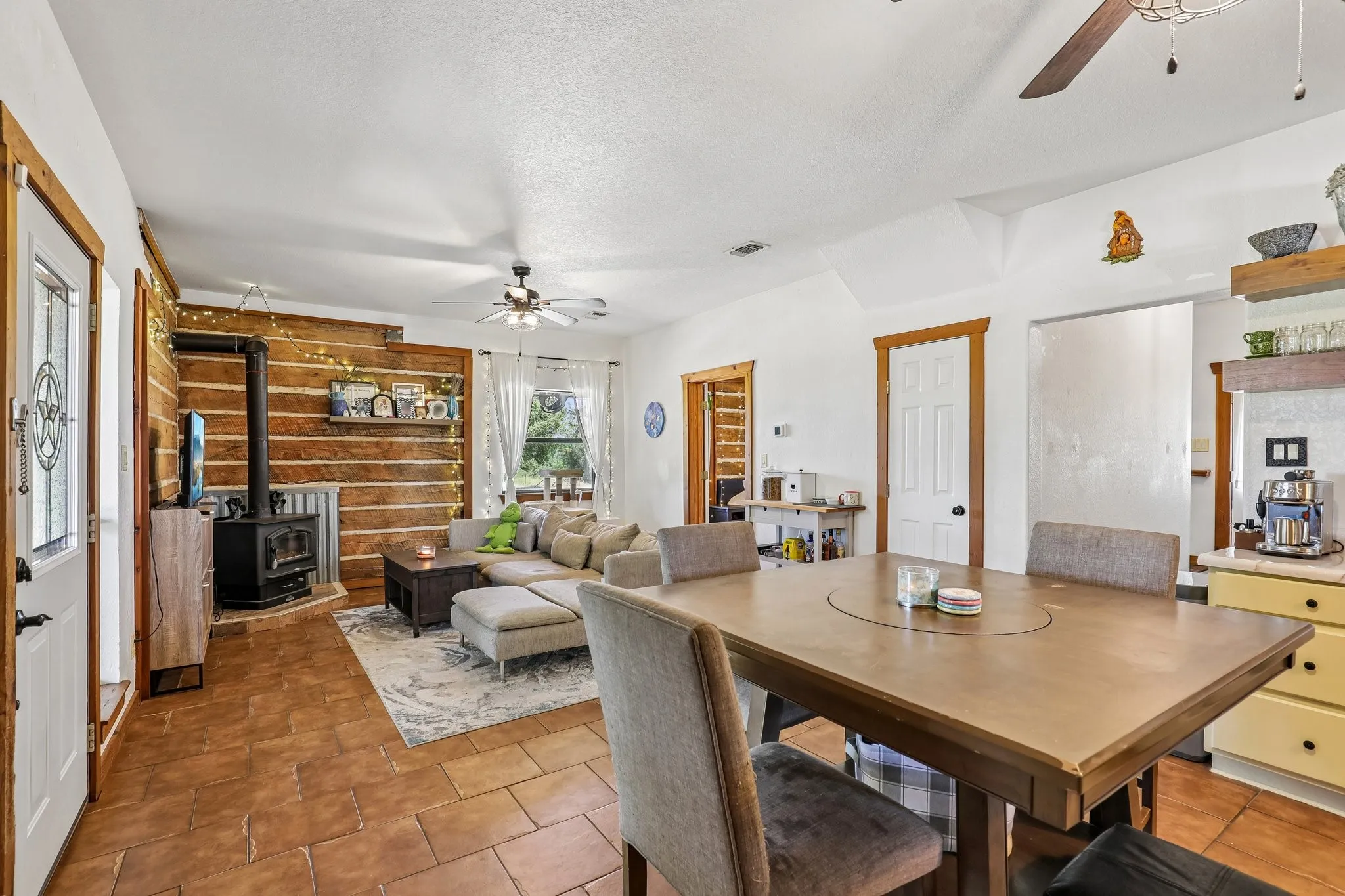 Open concept living area featuring a log wall accent, a wood-burning stove, and terracotta-style tile flooring