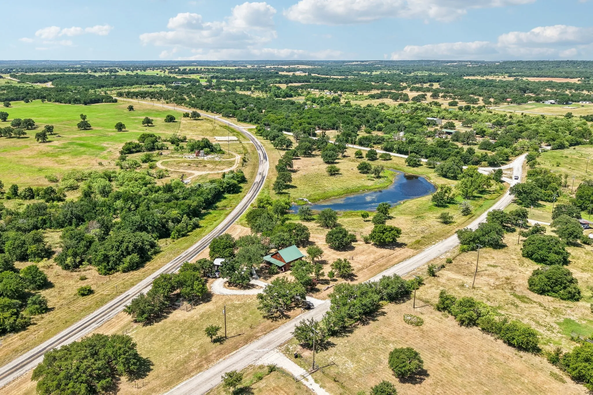 Expansive aerial view of a rural landscape featuring a natural pond, a single-story residence with a green roof, a private gravel road, and surrounding mature trees