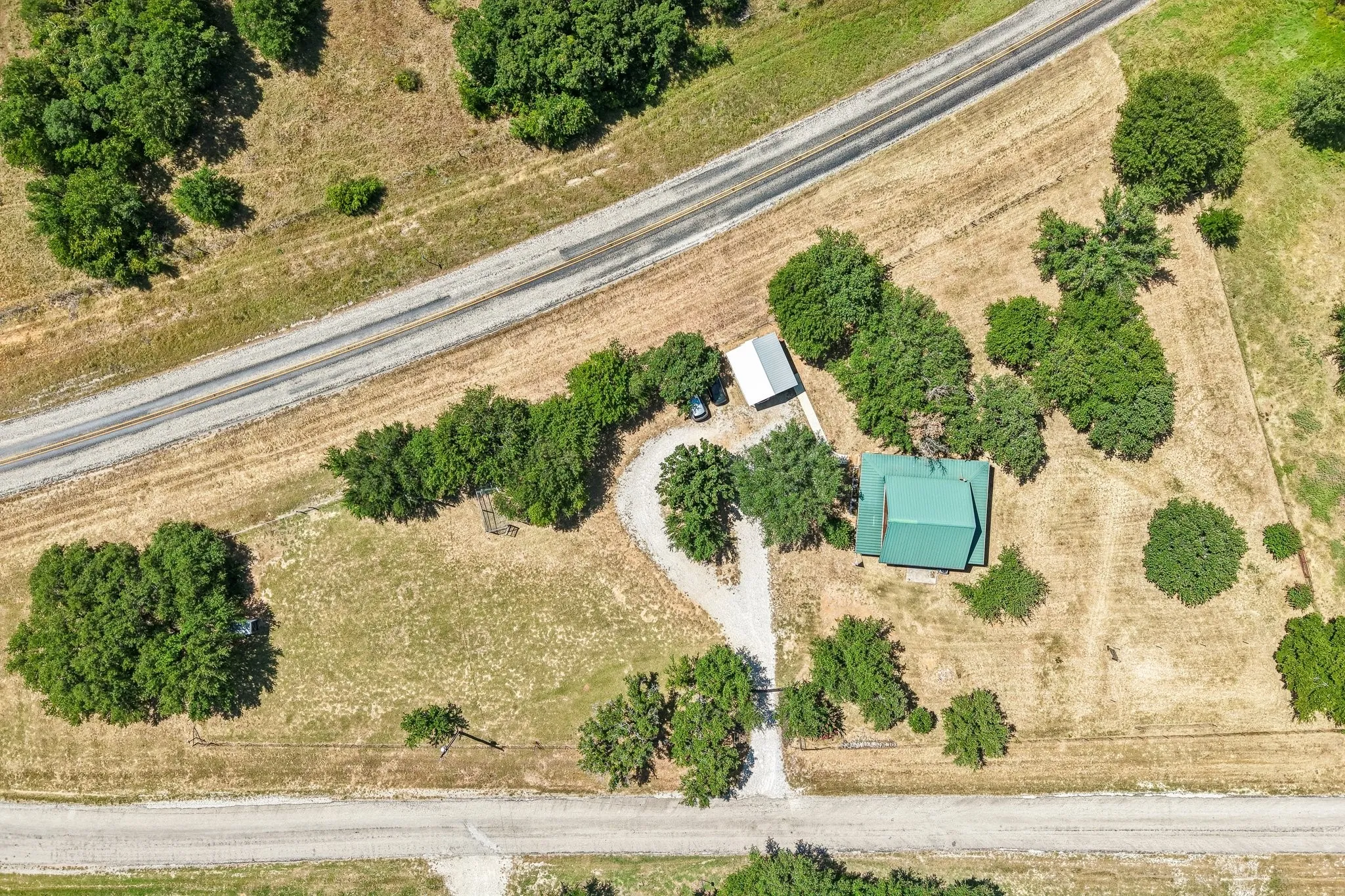 Aerial property overview featuring a green metal roof structure, unpaved drive, and surrounding natural landscape