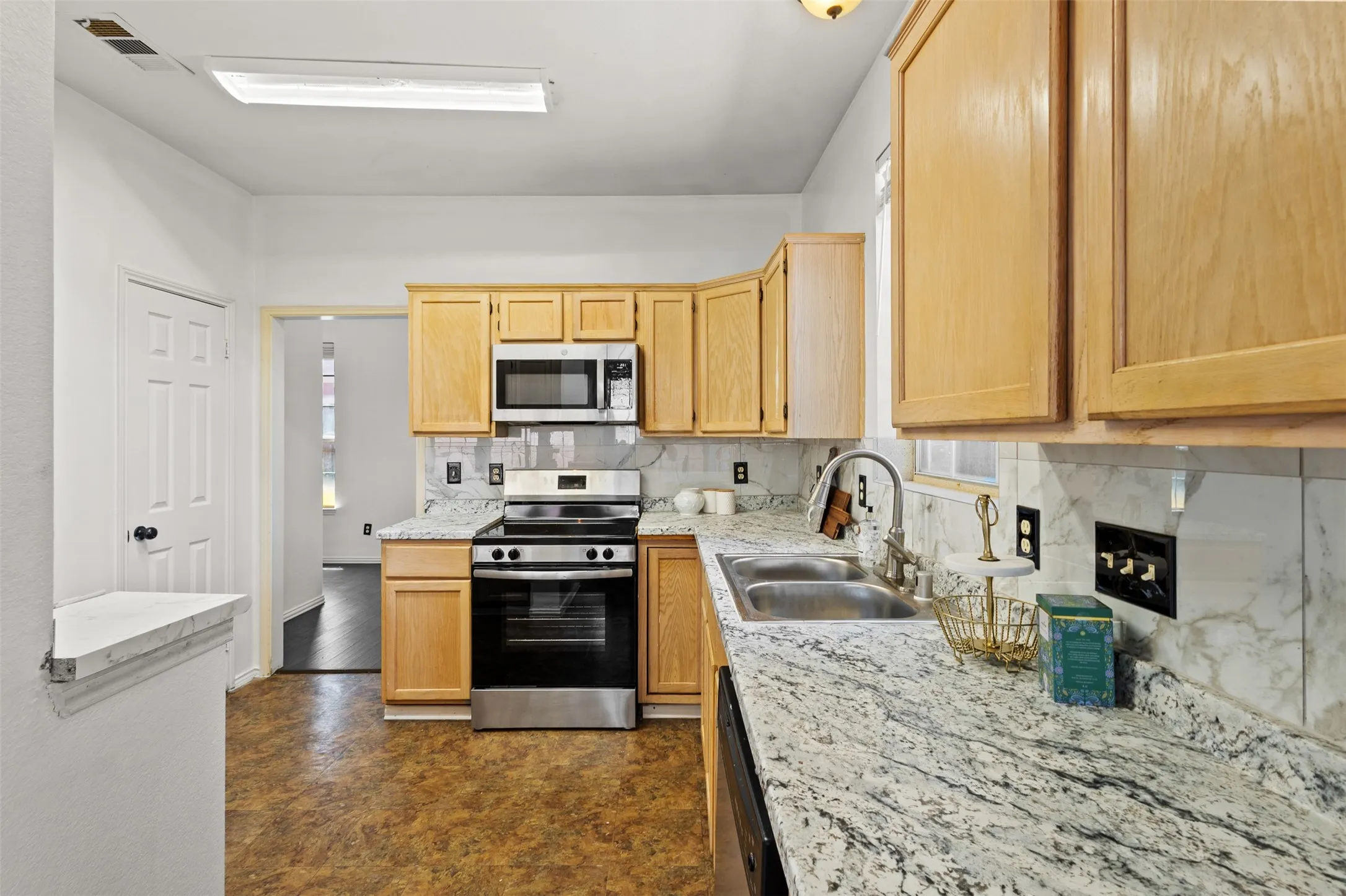 Kitchen featuring light wood cabinetry, stainless steel appliances, a double basin sink, and granite-look countertops with a matching backsplash