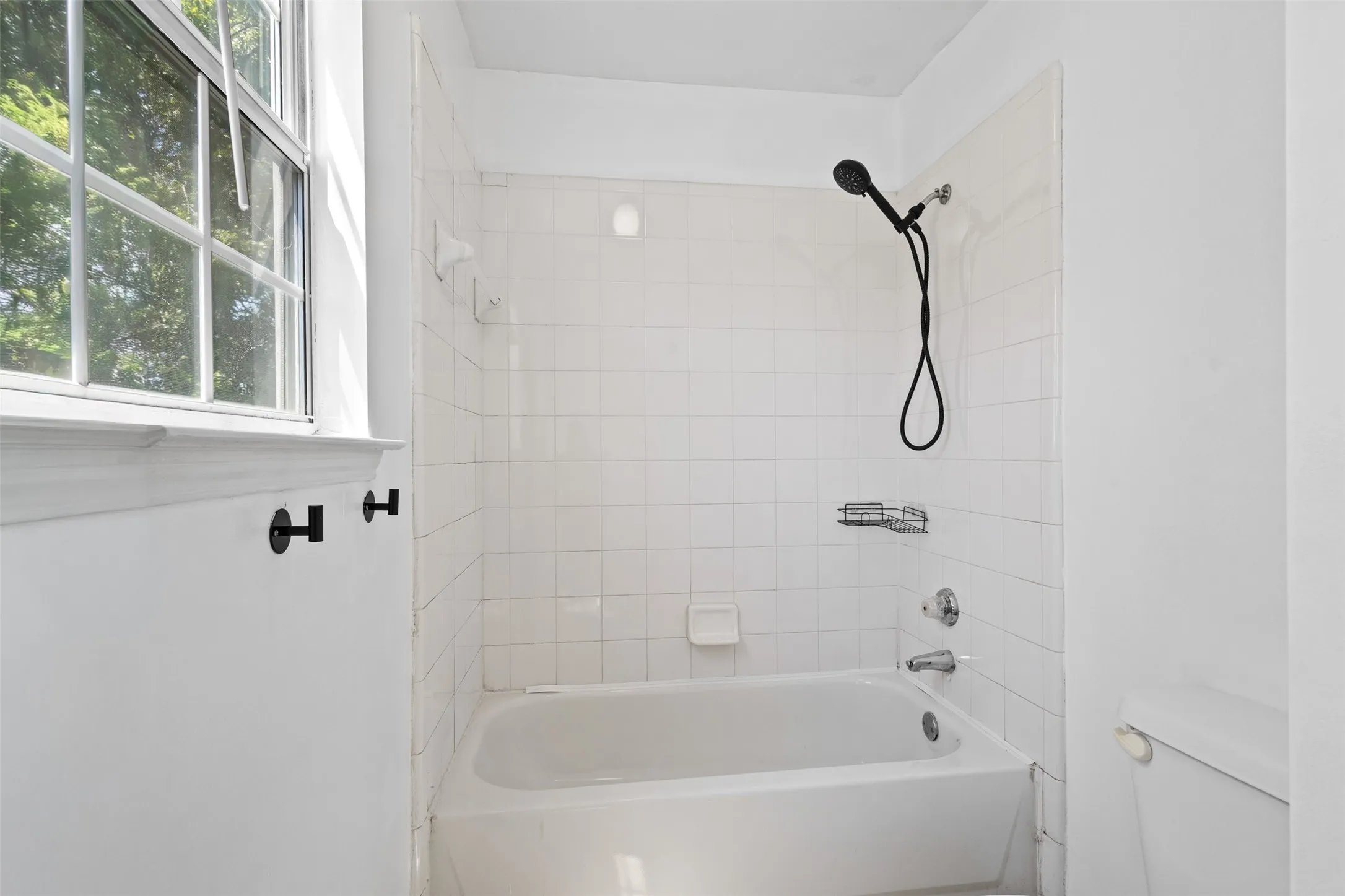 Bathroom featuring a white bathtub with a tile surround, a black handheld showerhead, and a window with a white frame