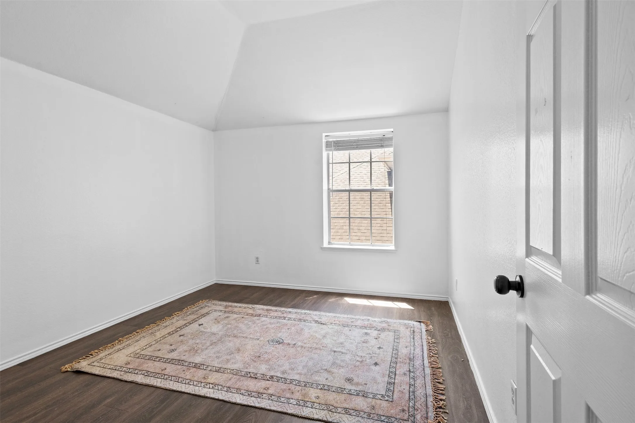 Vaulted ceiling room with wood-finish flooring, white baseboards, and a paneled white door with a black knob