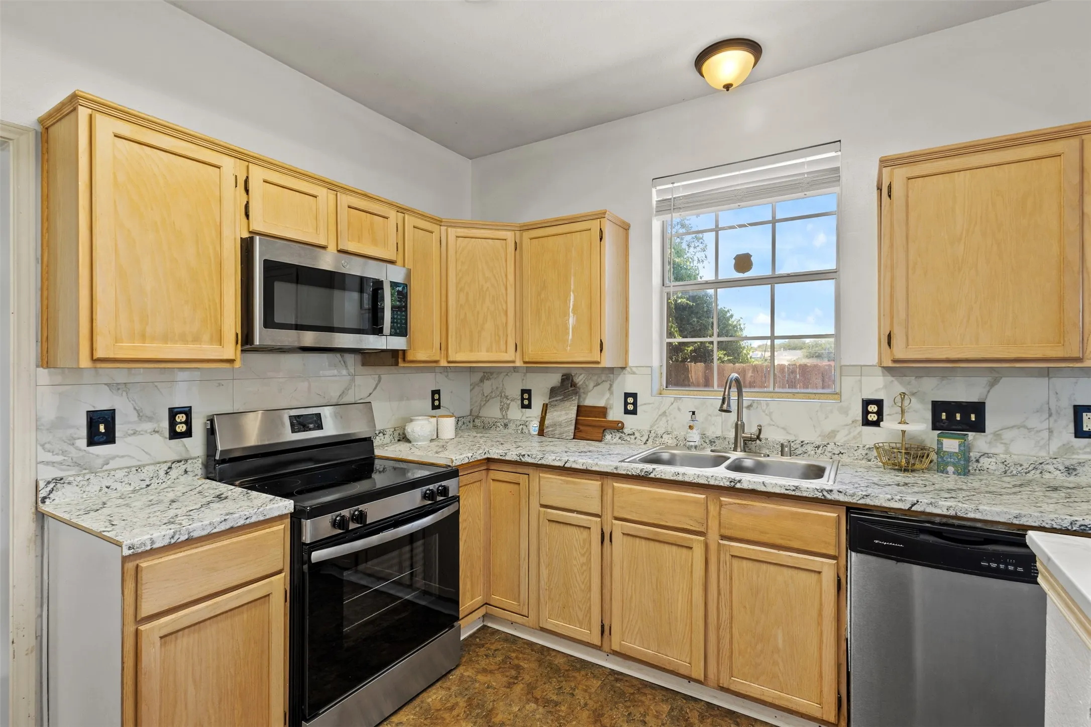 Kitchen featuring wood cabinetry, light-colored countertops with a white and gray pattern, stainless steel appliances, a double basin sink with a gooseneck faucet, and a window with blinds