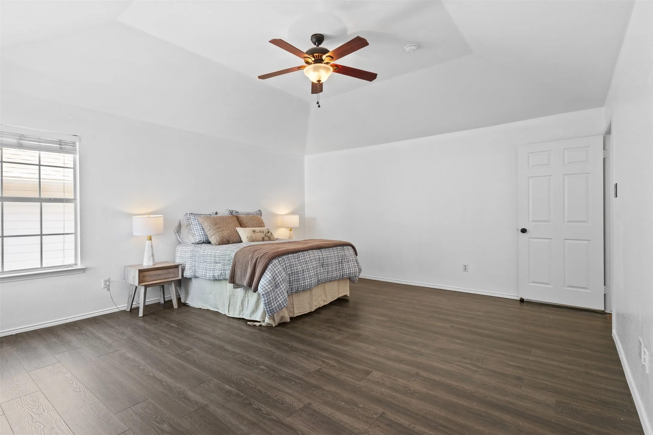 Spacious room featuring vaulted ceilings, dark wood-finish flooring, a ceiling fan with integrated lighting, and a paneled interior door