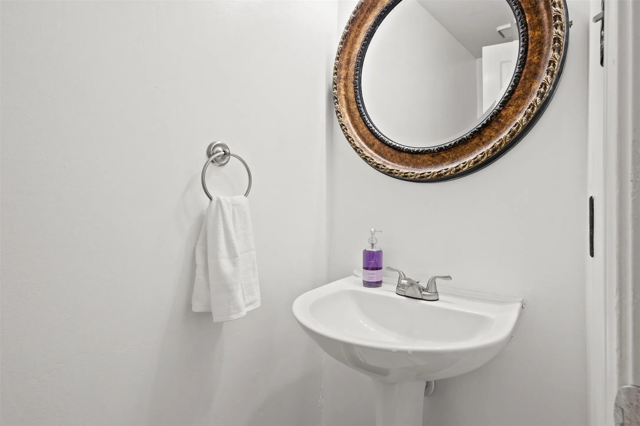 Pedestal sink with a chrome faucet, complemented by an ornate circular mirror and a wall-mounted towel ring