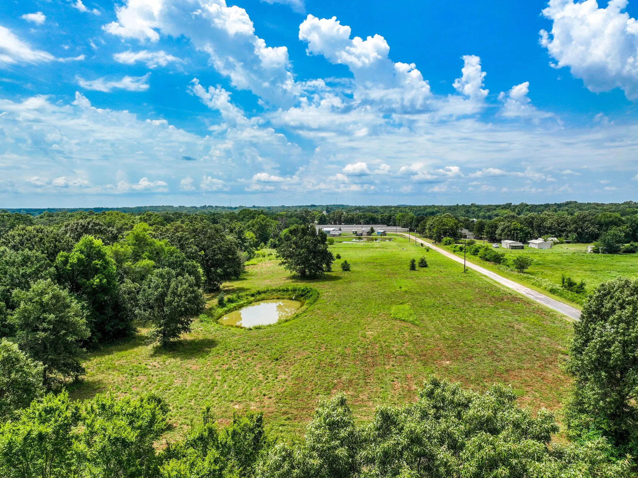 Expansive green acreage featuring a serene pond, mature trees, and a paved road running through the property