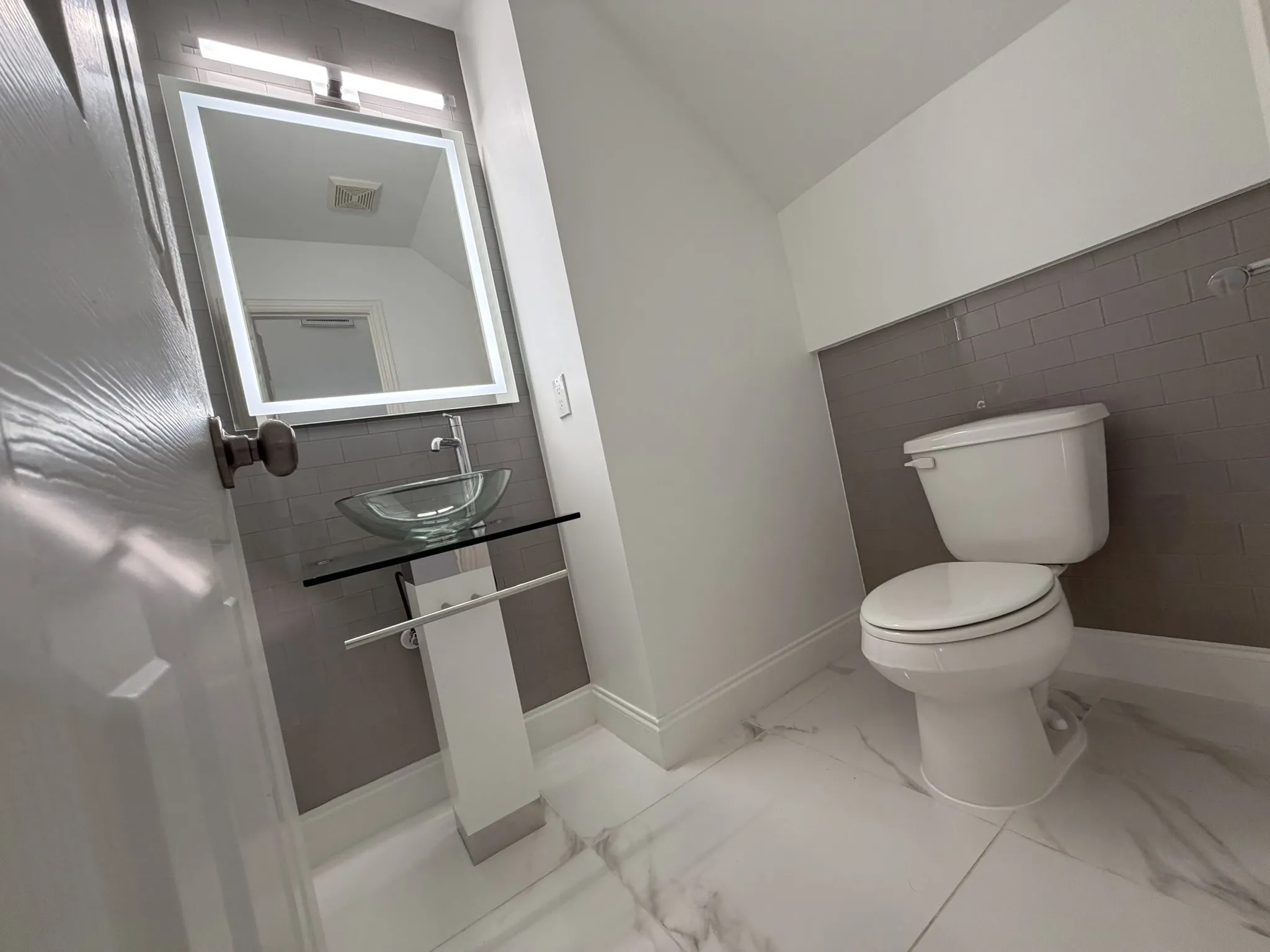 Powder room featuring a sleek illuminated mirror, clear vessel sink, pedestal-style vanity with integrated towel bar, and a grey subway tile accent wall
