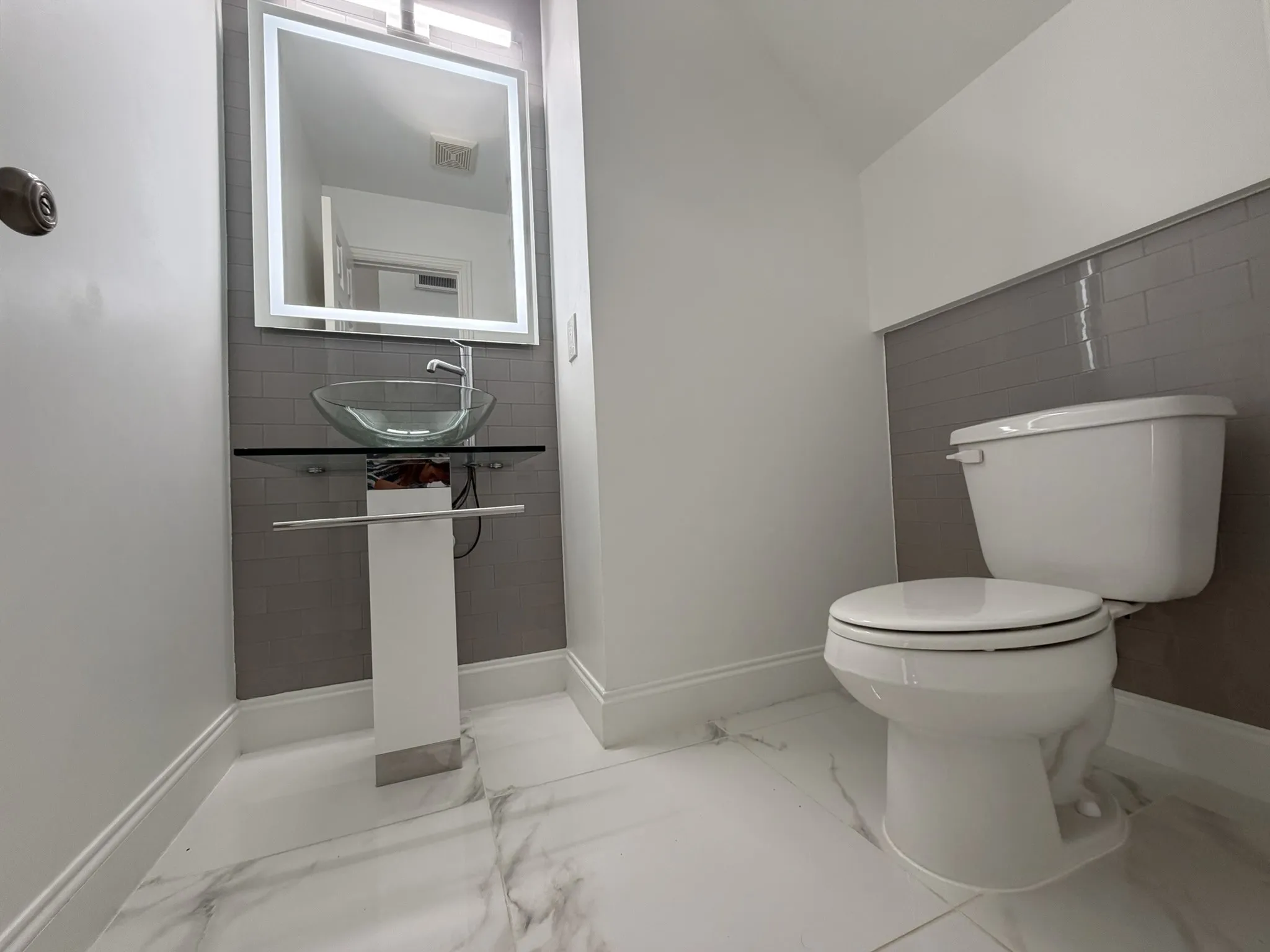 Contemporary powder room featuring a vessel sink with a glass countertop, an illuminated mirror, subway tile accent wall, and marble-look tile flooring