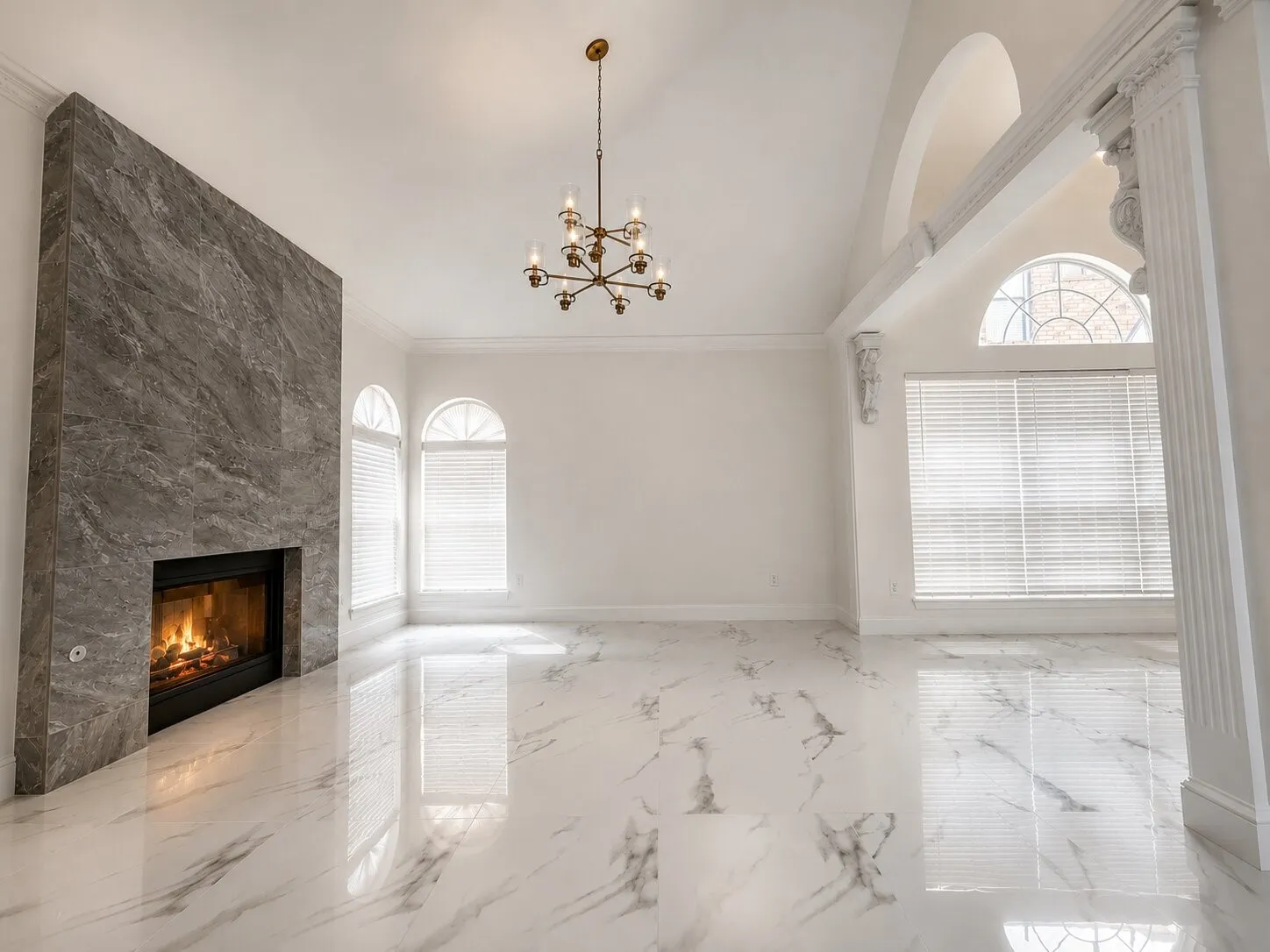 Spacious interior featuring a floor-to-ceiling stone-faced fireplace with a hearth, polished tile flooring, arched windows with blinds, a vaulted ceiling, and an ornate chandelier
