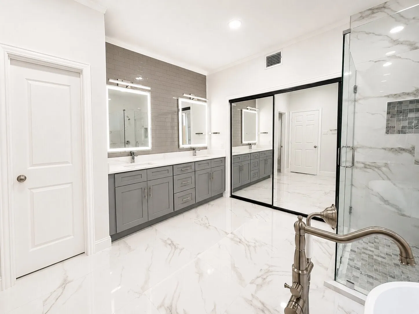 Spacious bathroom featuring white marble-look tile flooring, a dual vanity with gray cabinetry and white countertops, illuminated mirrors, and a glass-enclosed shower with white marble-look tile walls