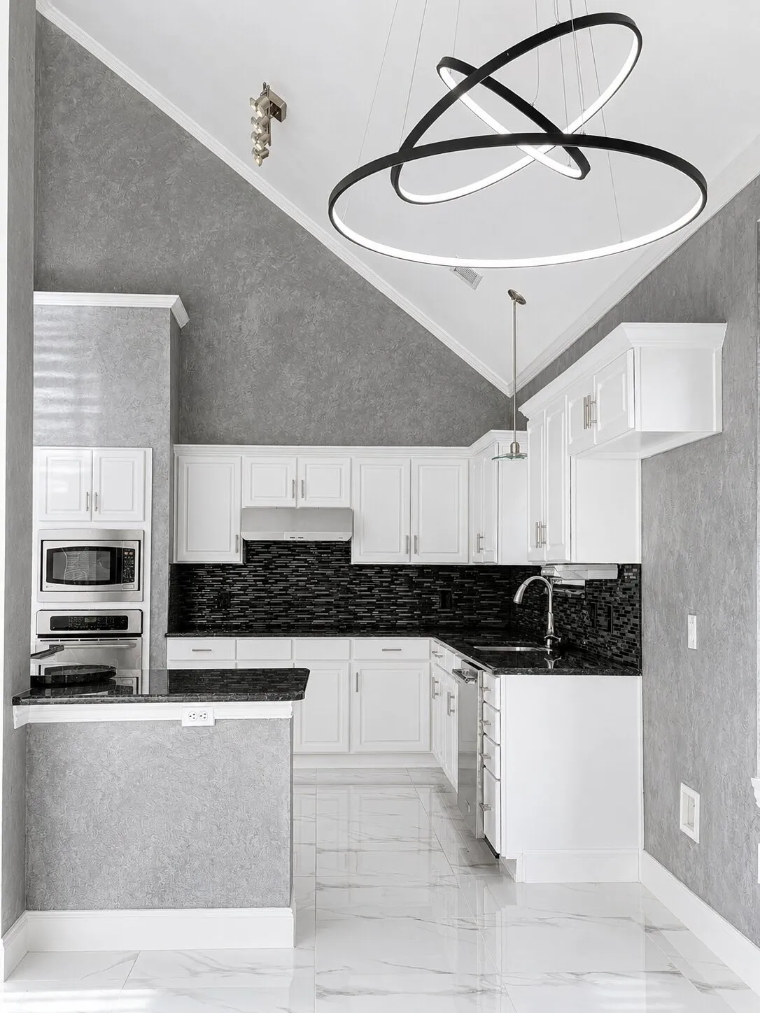 Modern kitchen featuring a striking geometric chandelier, vaulted ceiling, white cabinetry, dark countertops, and a full suite of stainless steel appliances