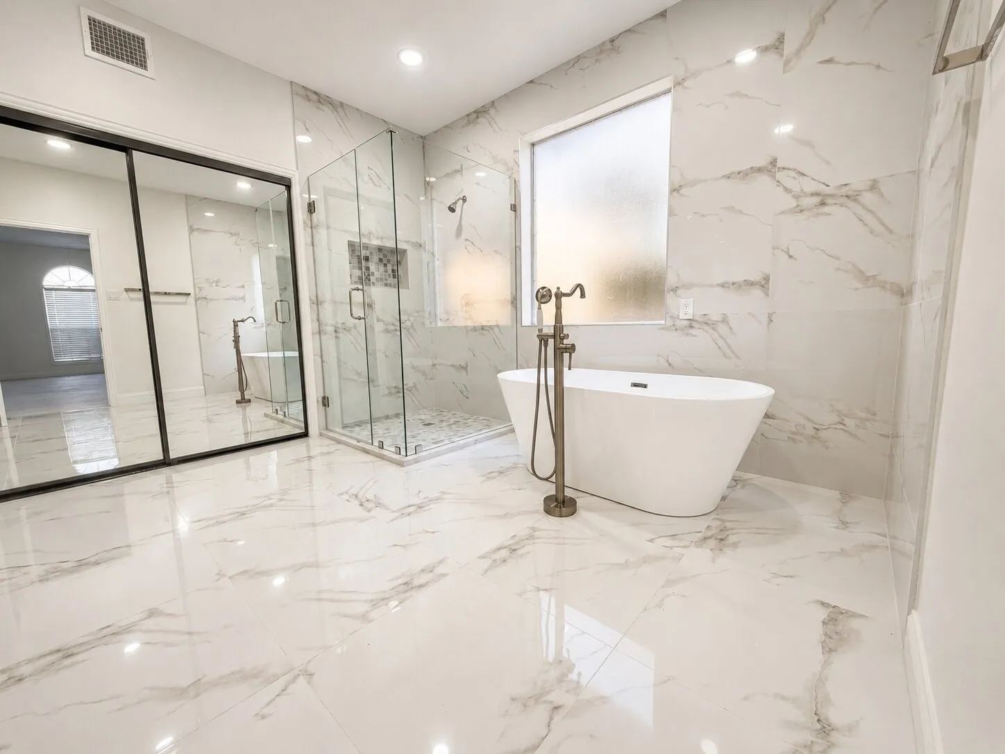 Spacious bathroom featuring large format marble-look tile flooring and walls, a freestanding soaking tub with bronze-finish floor-mounted faucet, and a frameless glass shower enclosure with a tiled niche