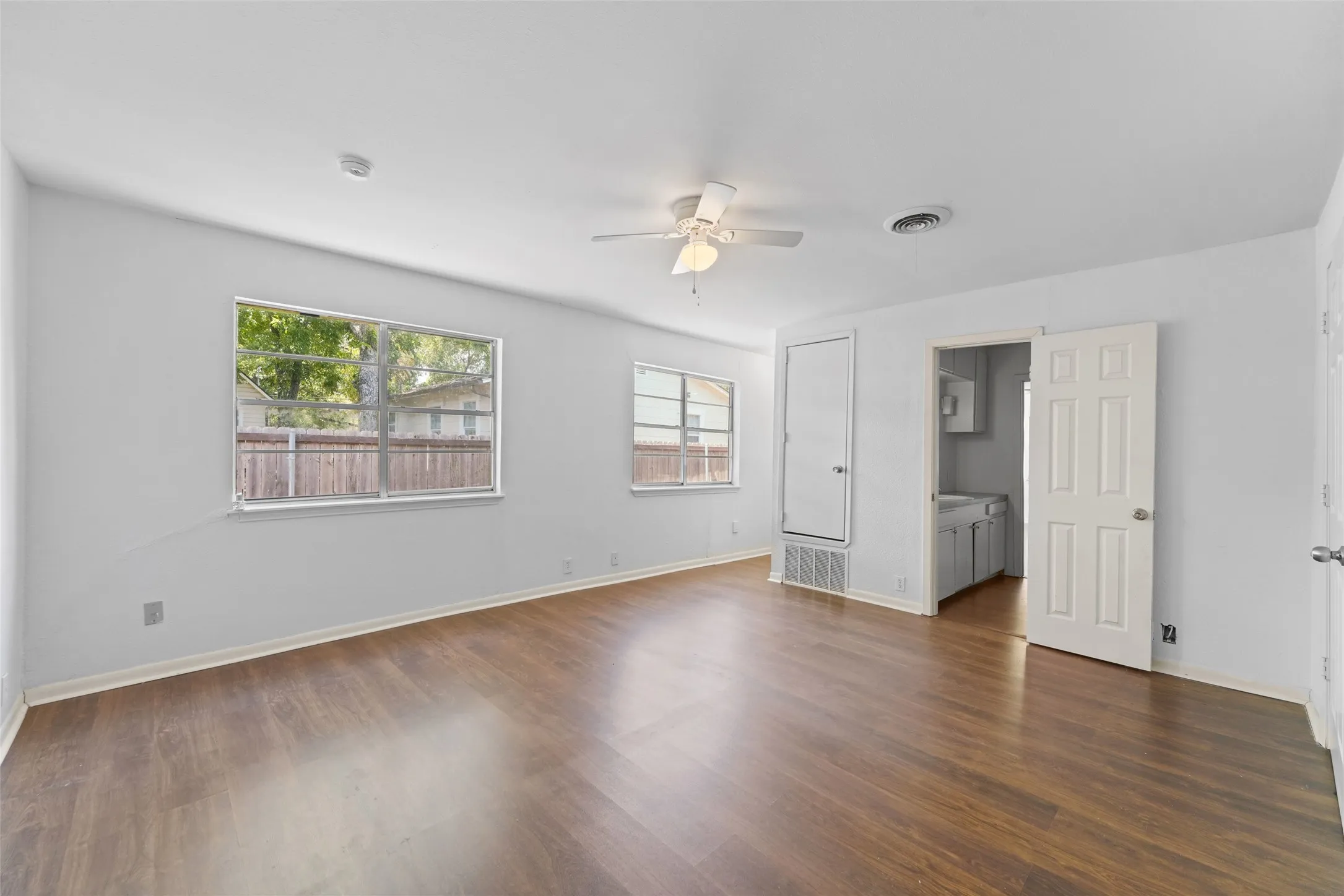 Wood-finish flooring throughout, white walls, multiple windows, ceiling fan with light, and a paneled interior door
