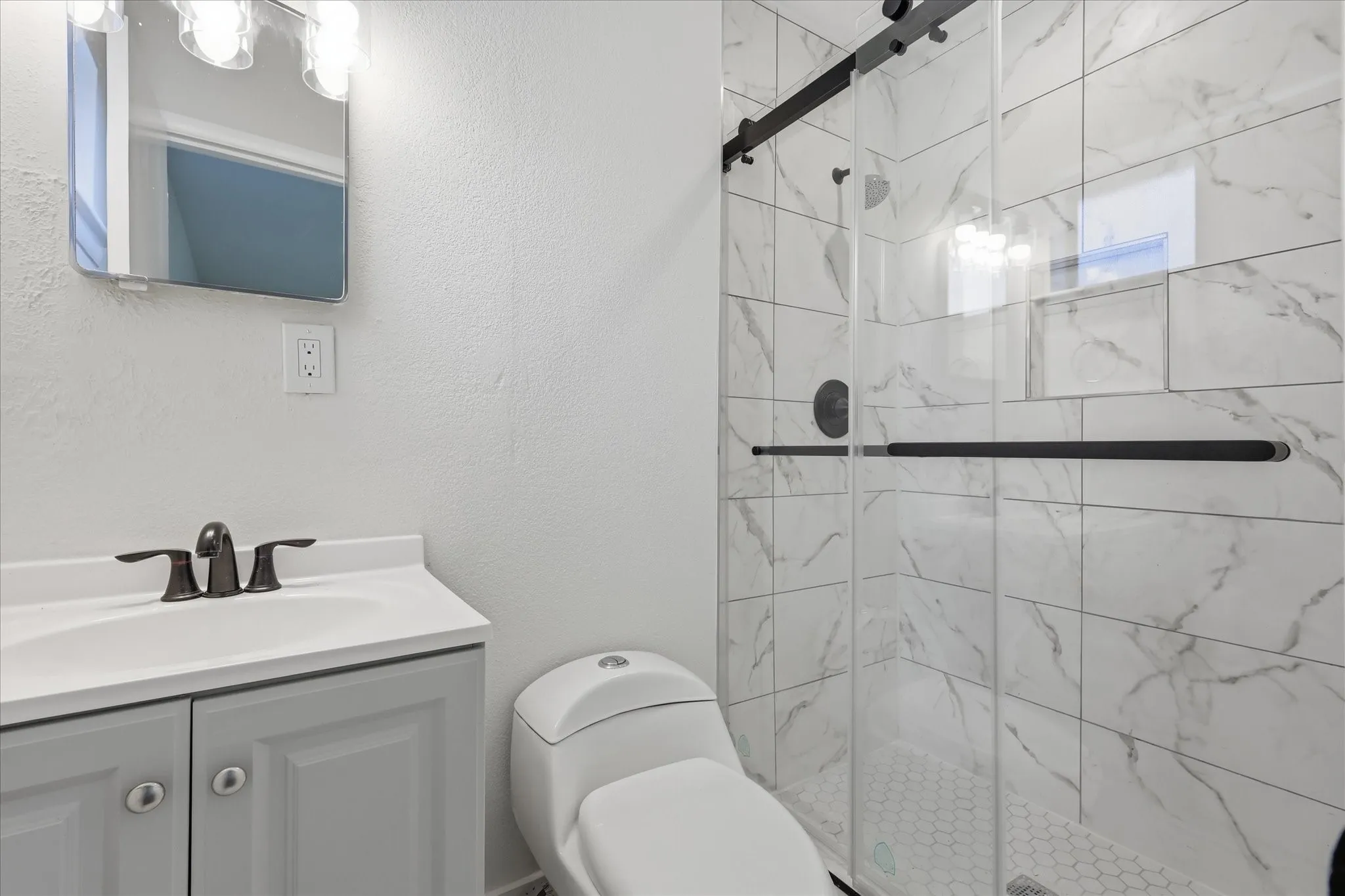 Bathroom featuring a glass-enclosed shower with large format marble-look tile, a white hexagonal tile shower floor, a light gray vanity with a white countertop and integrated sink, a dark bronze faucet, and a wall-mounted mirror with a vanity light