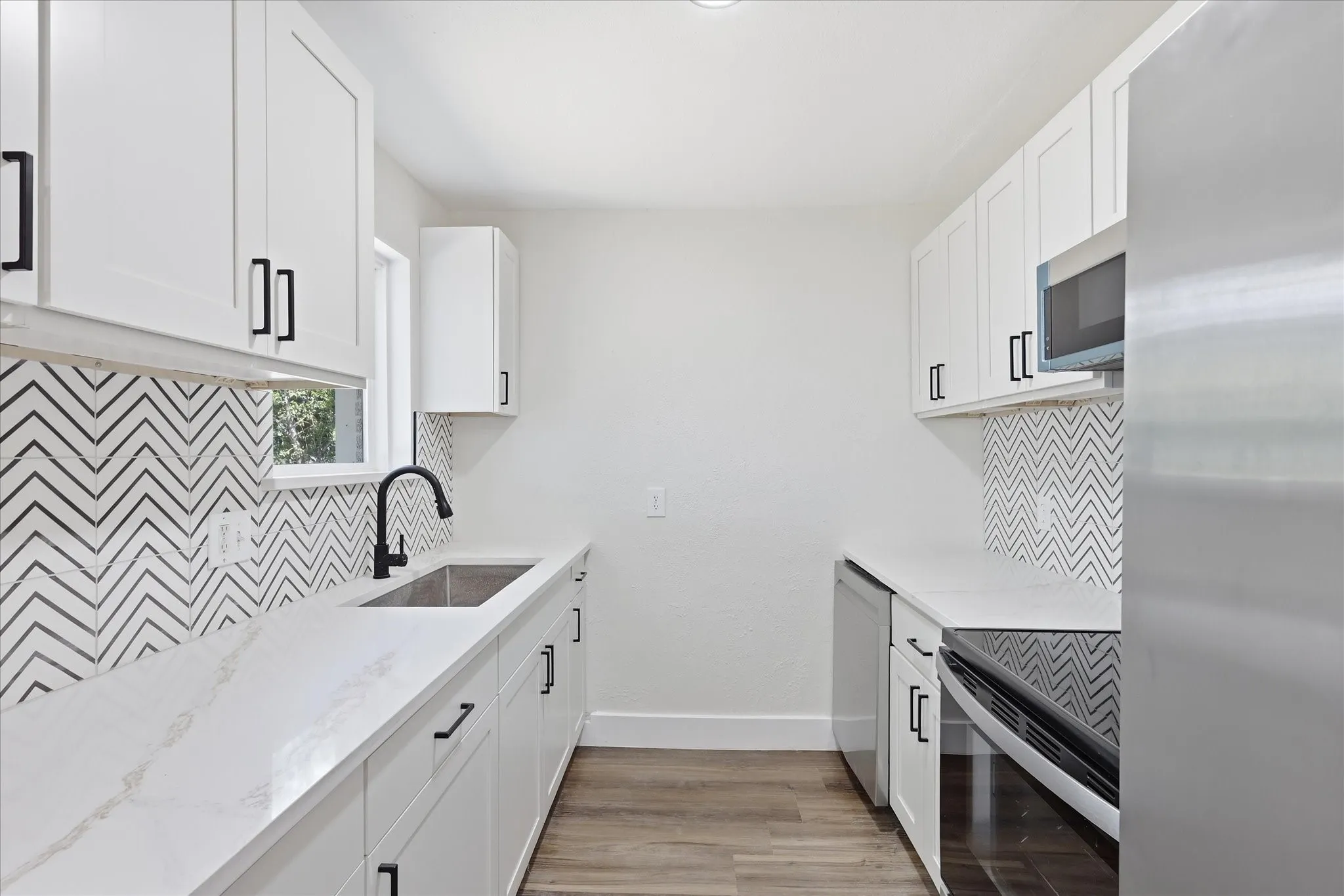 Modern kitchen featuring white shaker cabinetry with matte black hardware, white quartz countertops, a chevron-patterned tile backsplash, and stainless steel appliances