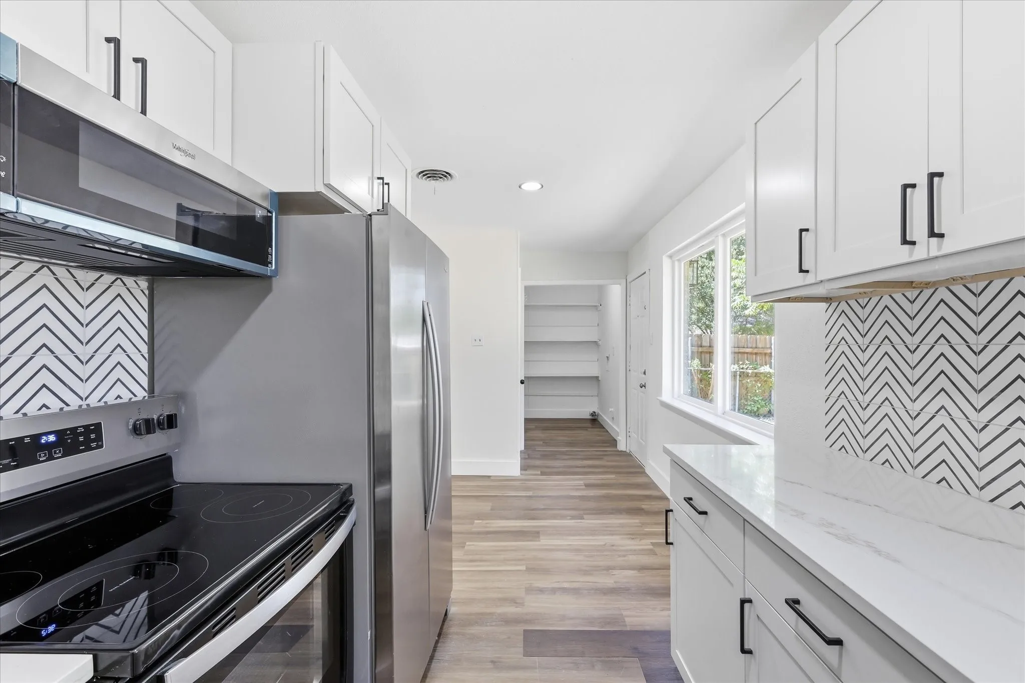 Contemporary kitchen featuring white shaker cabinetry with black hardware, stainless steel appliances, a chevron pattern tile backsplash, light-toned wood-finish flooring, and a window providing natural light