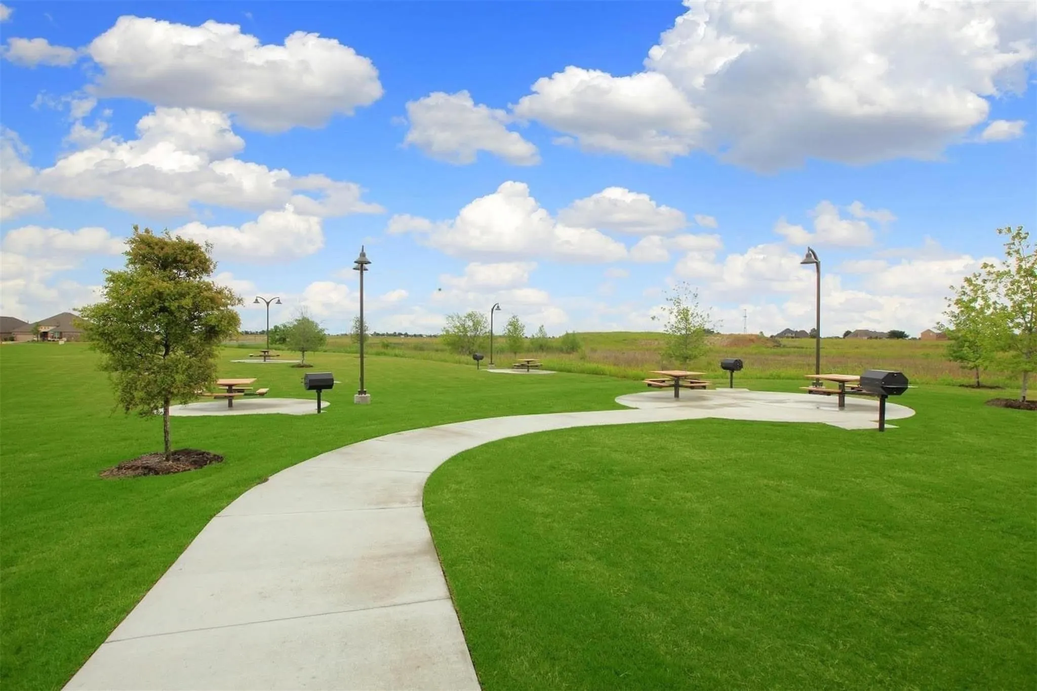 Expansive green space featuring a concrete walking path, mature trees, designated picnic areas with tables, and built-in grilling stations