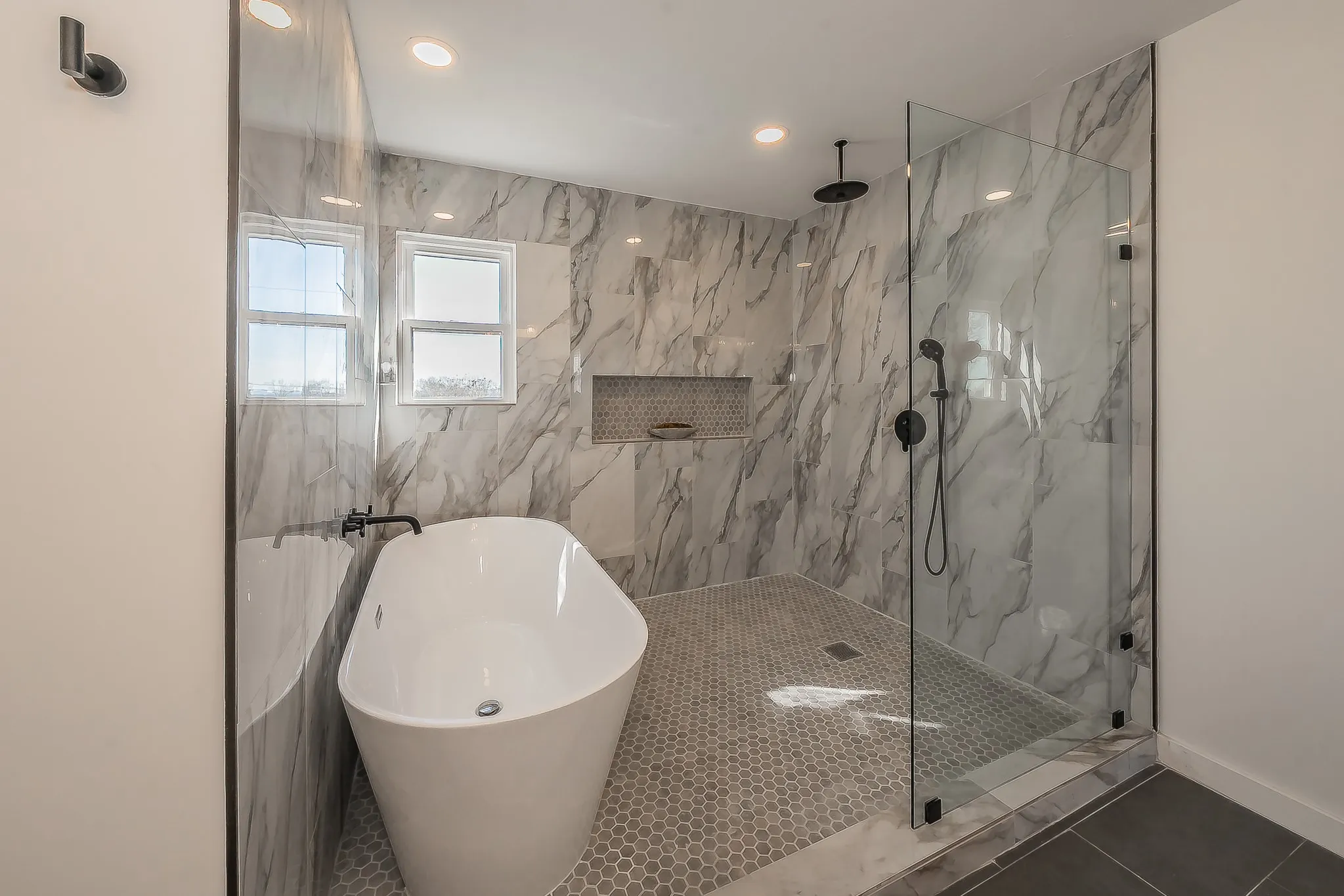 Contemporary bathroom featuring a freestanding tub, glass-enclosed shower with matte black fixtures, marble-finish wall tiling, recessed lighting, and hex-tile shower flooring