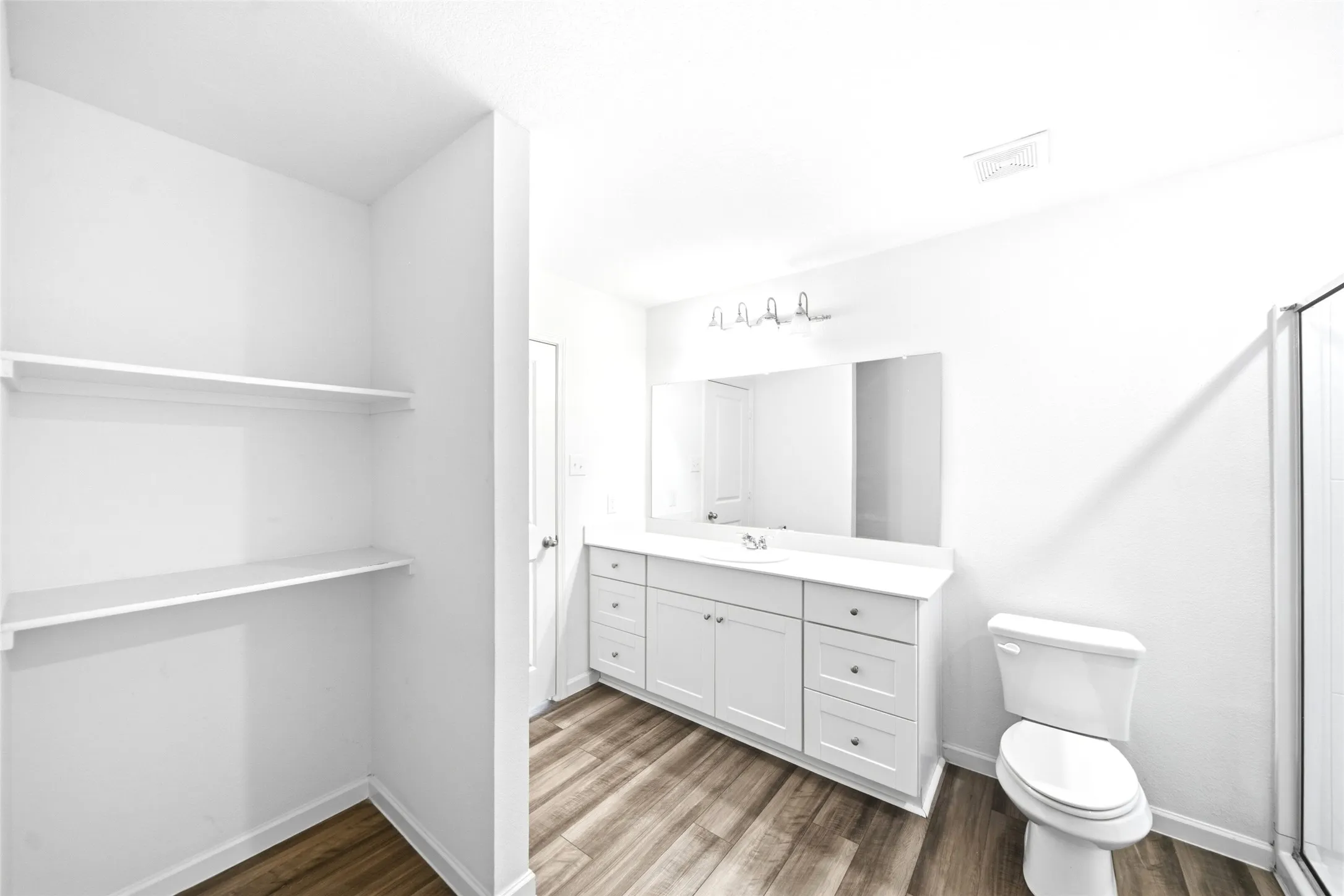 White vanity with a single sink, wood-finish flooring, built-in shelving, mirror, and a glass-enclosed shower