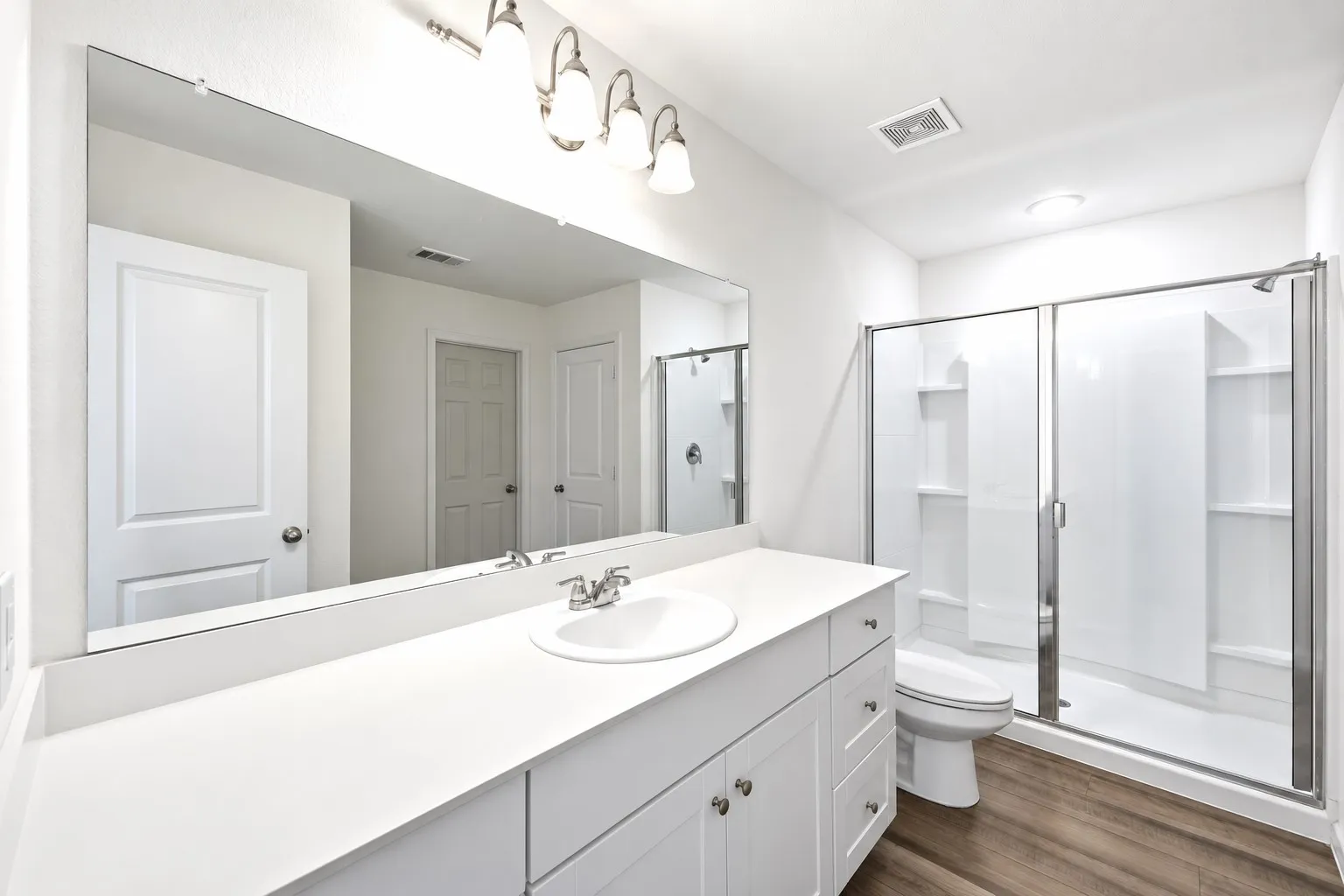 White vanity with integrated sink, expansive wall-mounted mirror, brushed nickel light fixture, glass-enclosed shower, and wood-finish flooring