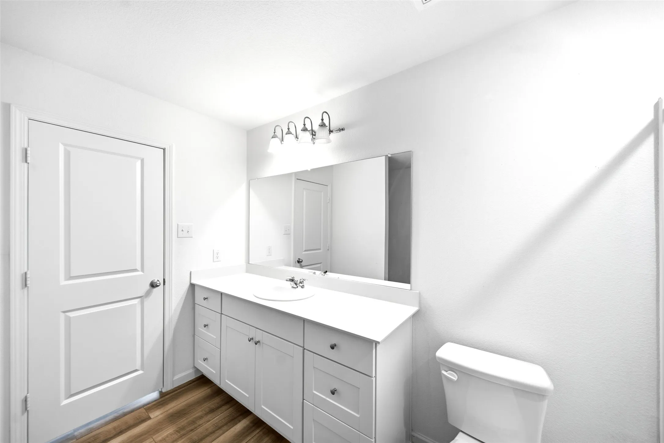 White vanity with integrated sink, rectangular wall mirror, four-light vanity fixture, white toilet, and wood-finish flooring
