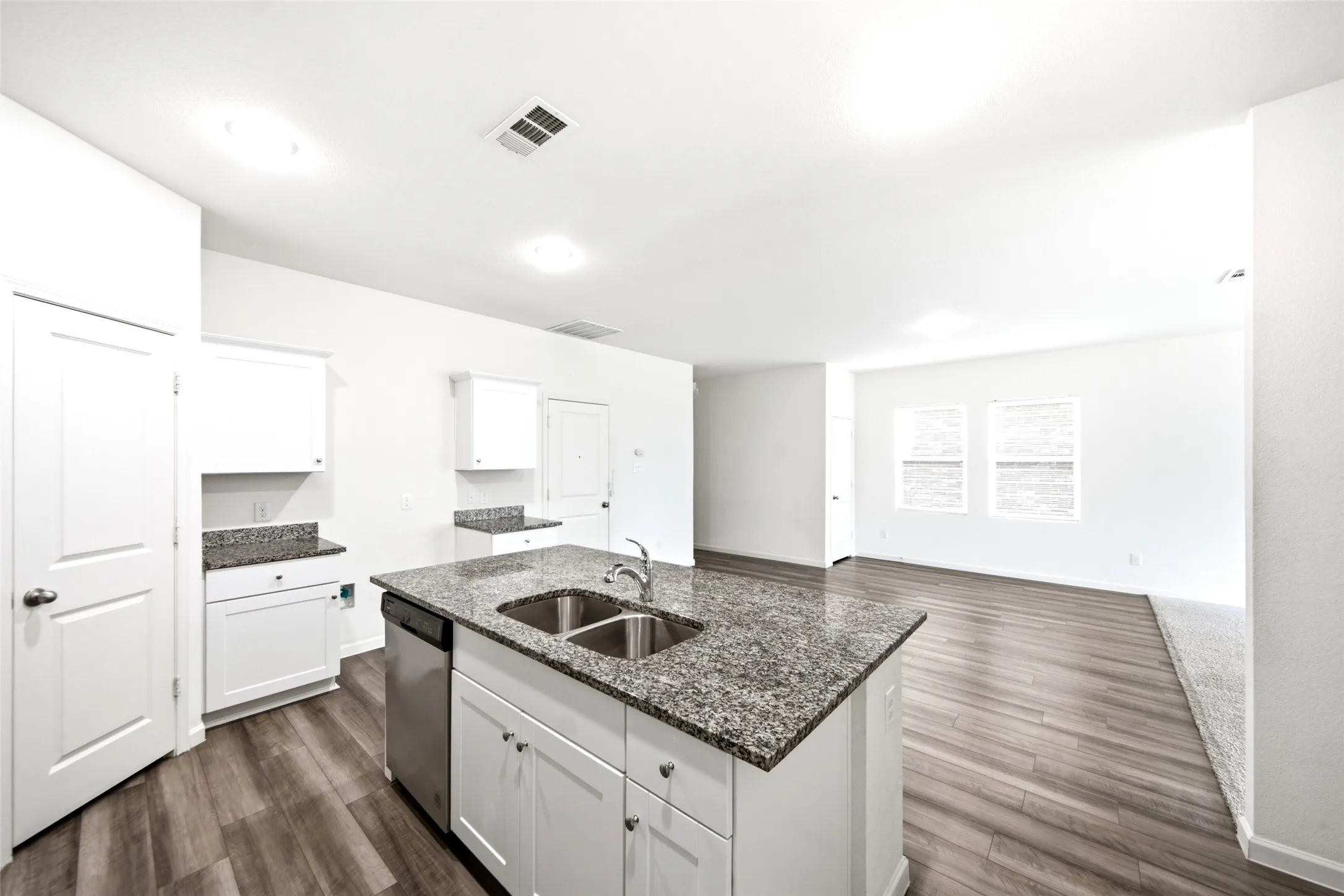 Kitchen island with dual stainless steel sink and granite countertop, white cabinetry with dark hardware, stainless steel dishwasher, and wood-finish flooring