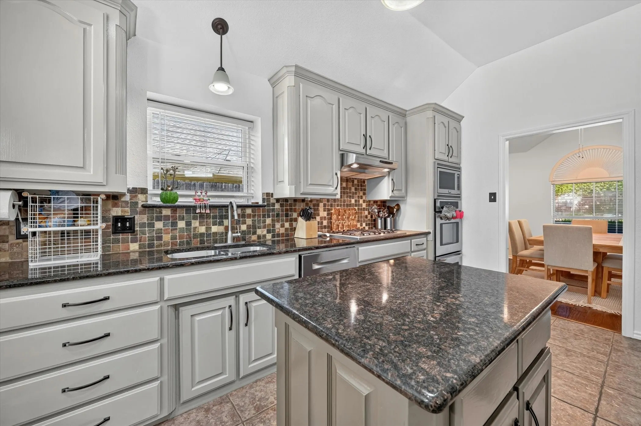 Kitchen featuring granite countertops, wood-finish cabinetry, tile backsplash, stainless steel appliances, and a vaulted ceiling