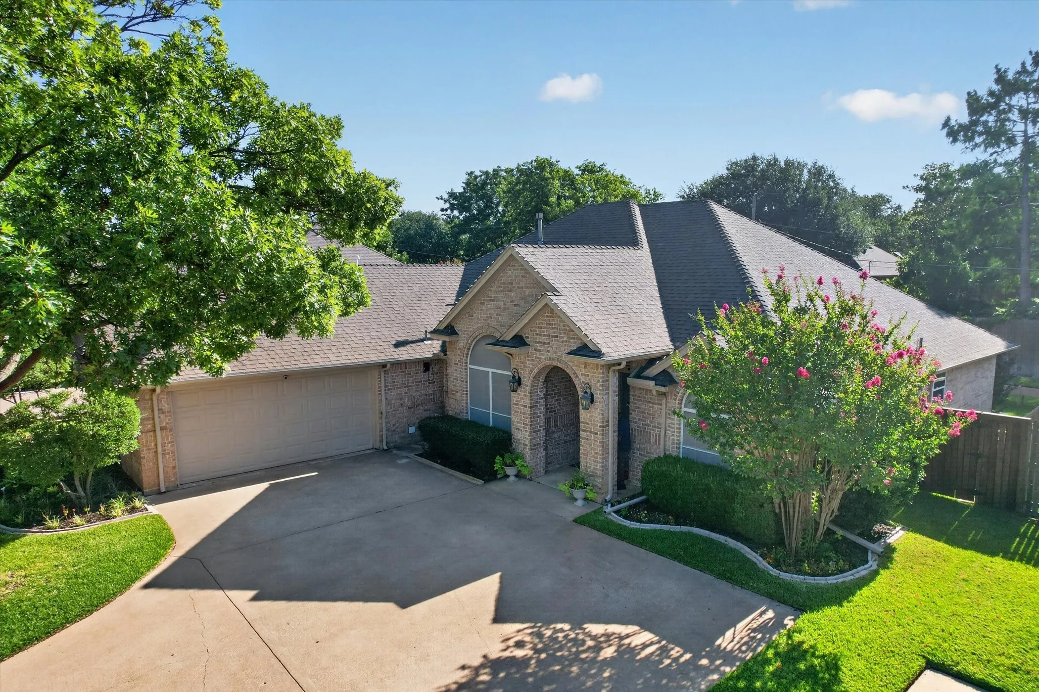 Brick facade residence featuring a prominent arched window, a dark shingle roof, mature trees, and a well-kept lawn