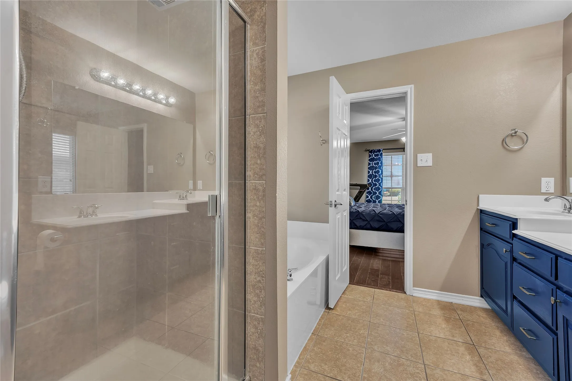 Primary bathroom featuring a spacious walk-in shower with glass enclosure and tile surround, dual vanities with blue cabinetry, ceramic tile flooring, and abundant lighting above the mirror