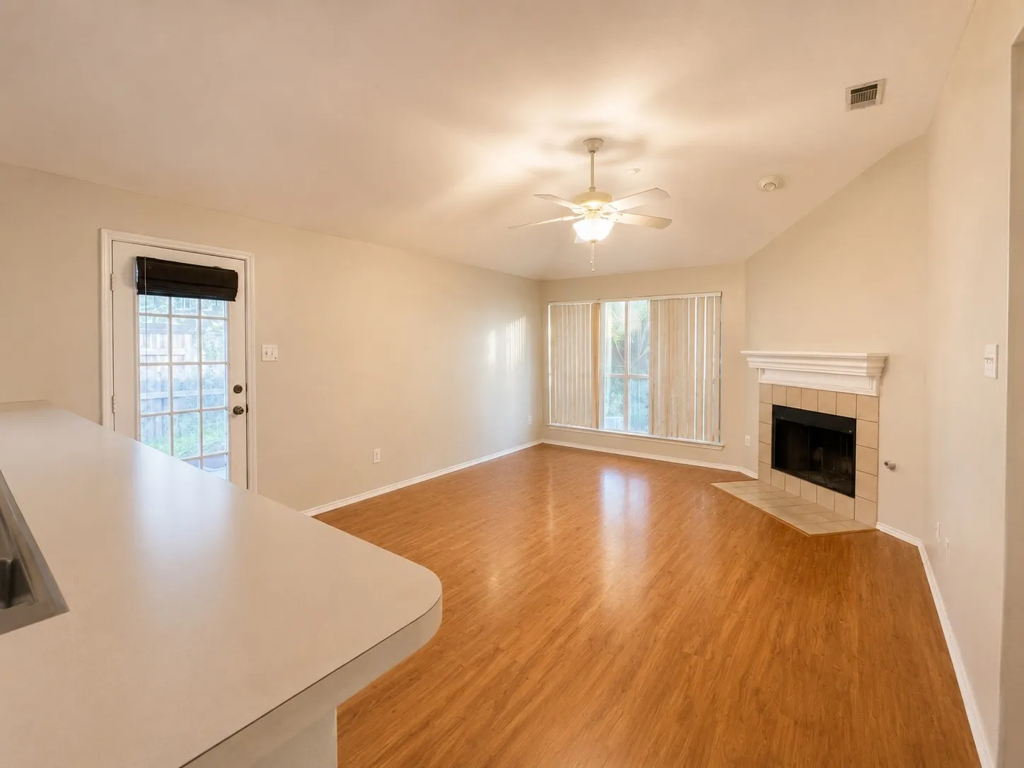 Spacious living area featuring wood-finish flooring, a tiled fireplace with mantel, a ceiling fan with integrated lighting, and multiple windows with blinds