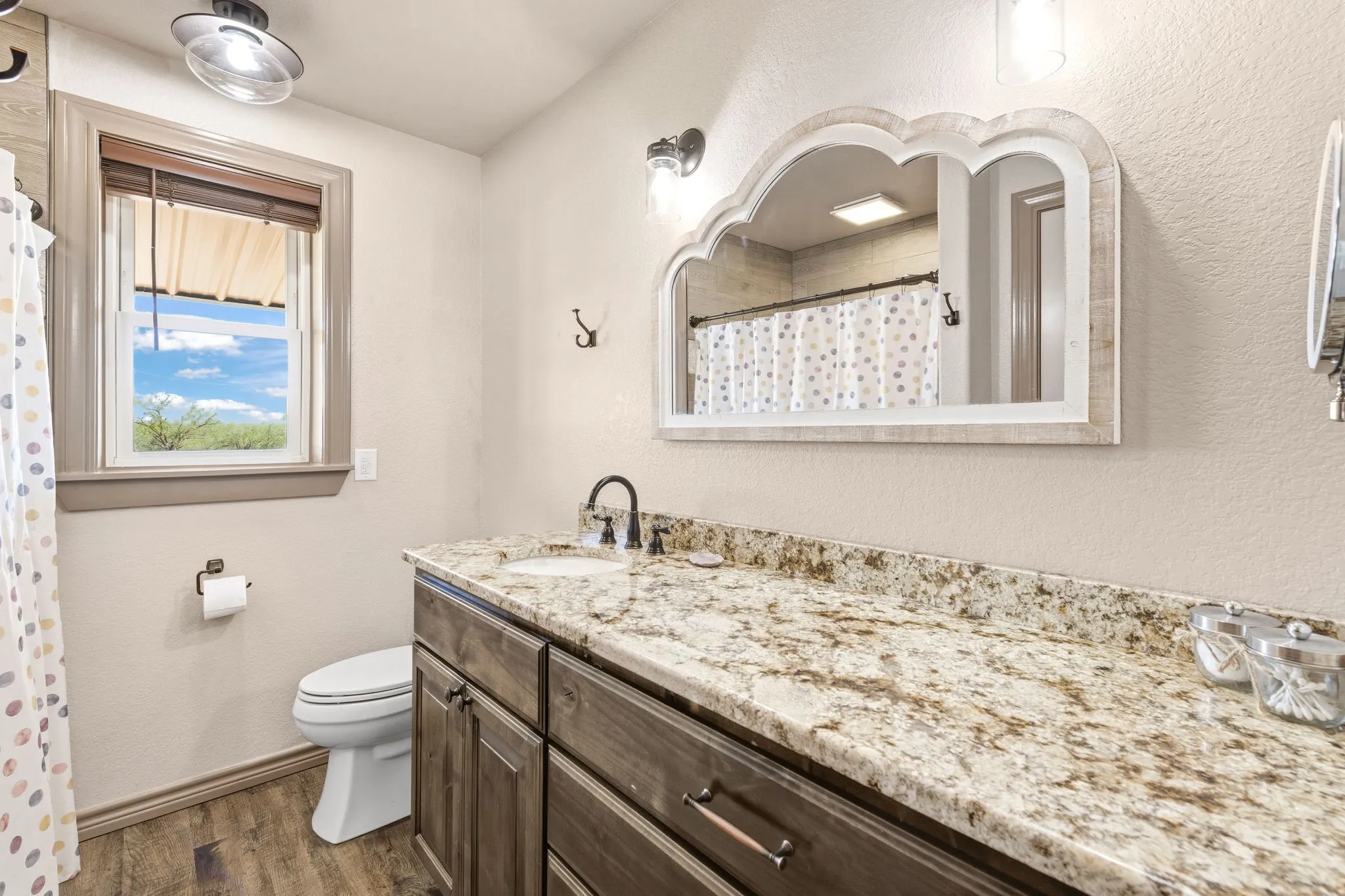 Guest bathroom offers a long granite countertop with plenty of space to spread out, arched mirror for character, LVP flooring and a standing shower plus a window bringing in natural light.