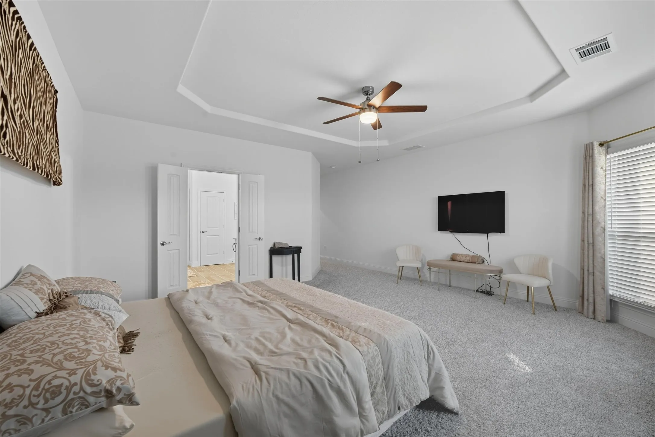 Spacious room featuring tray ceilings, neutral carpeting, a ceiling fan with integrated lighting, and multiple windows with blinds