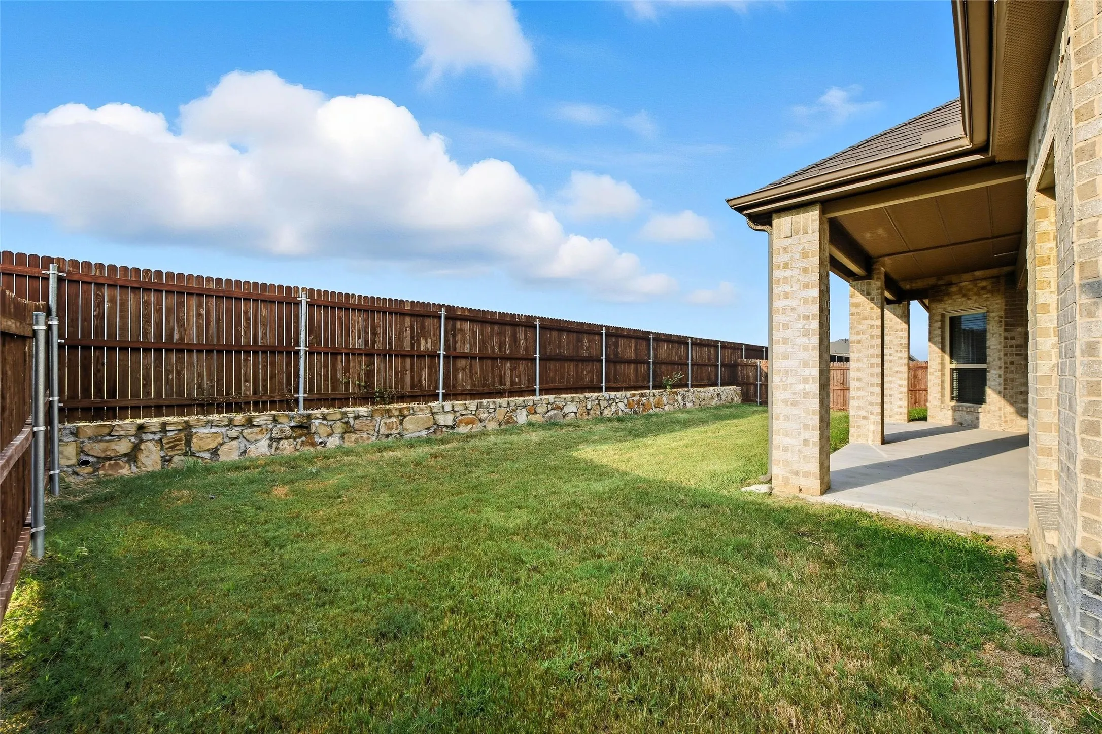Spacious backyard featuring a covered patio with brick columns, a manicured lawn, and a privacy fence with a stone retaining wall