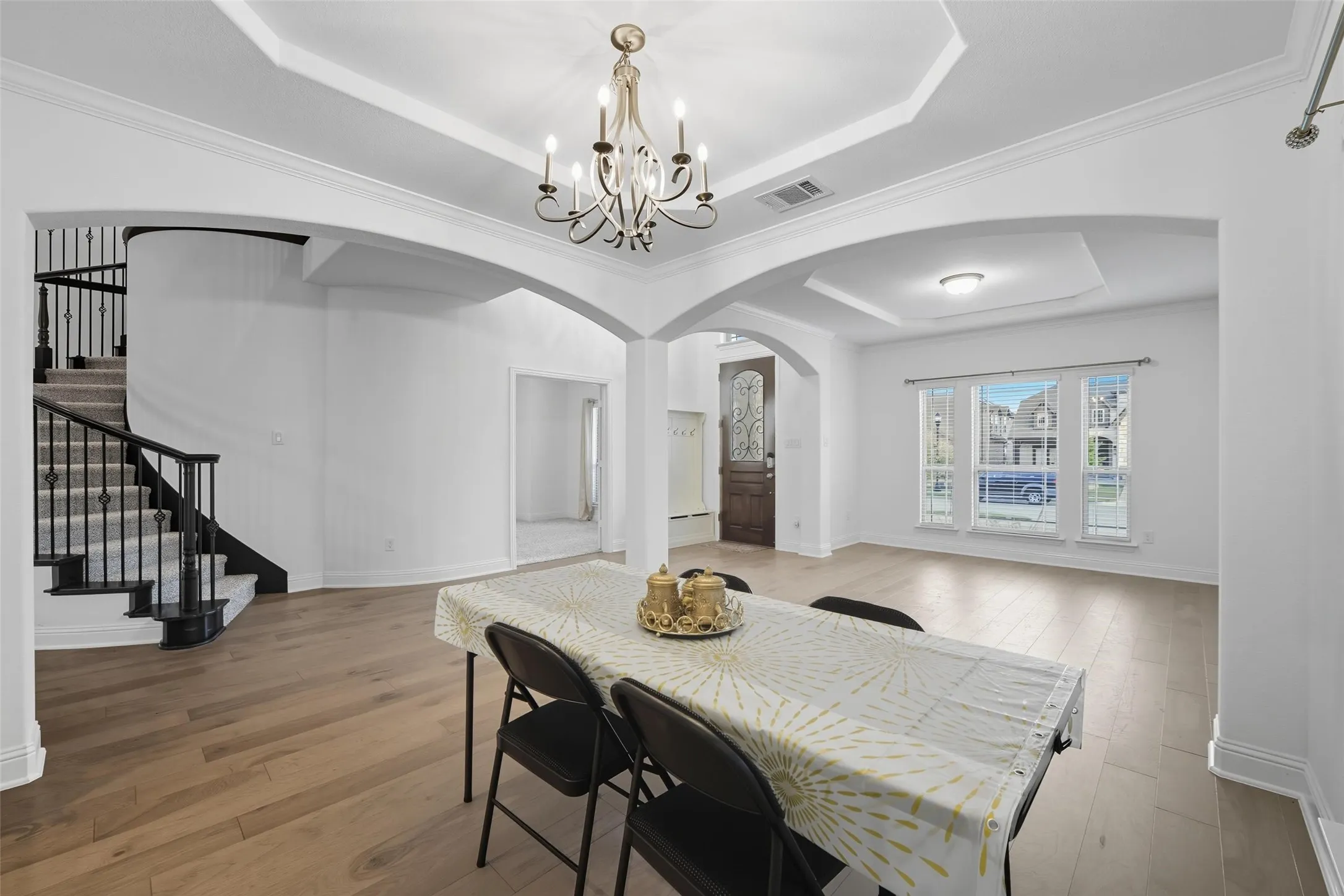 Open-concept interior featuring wood-finish flooring, a grand staircase with wrought iron balusters, arched doorways, a coffered ceiling with a chandelier, and a decorative front entry door