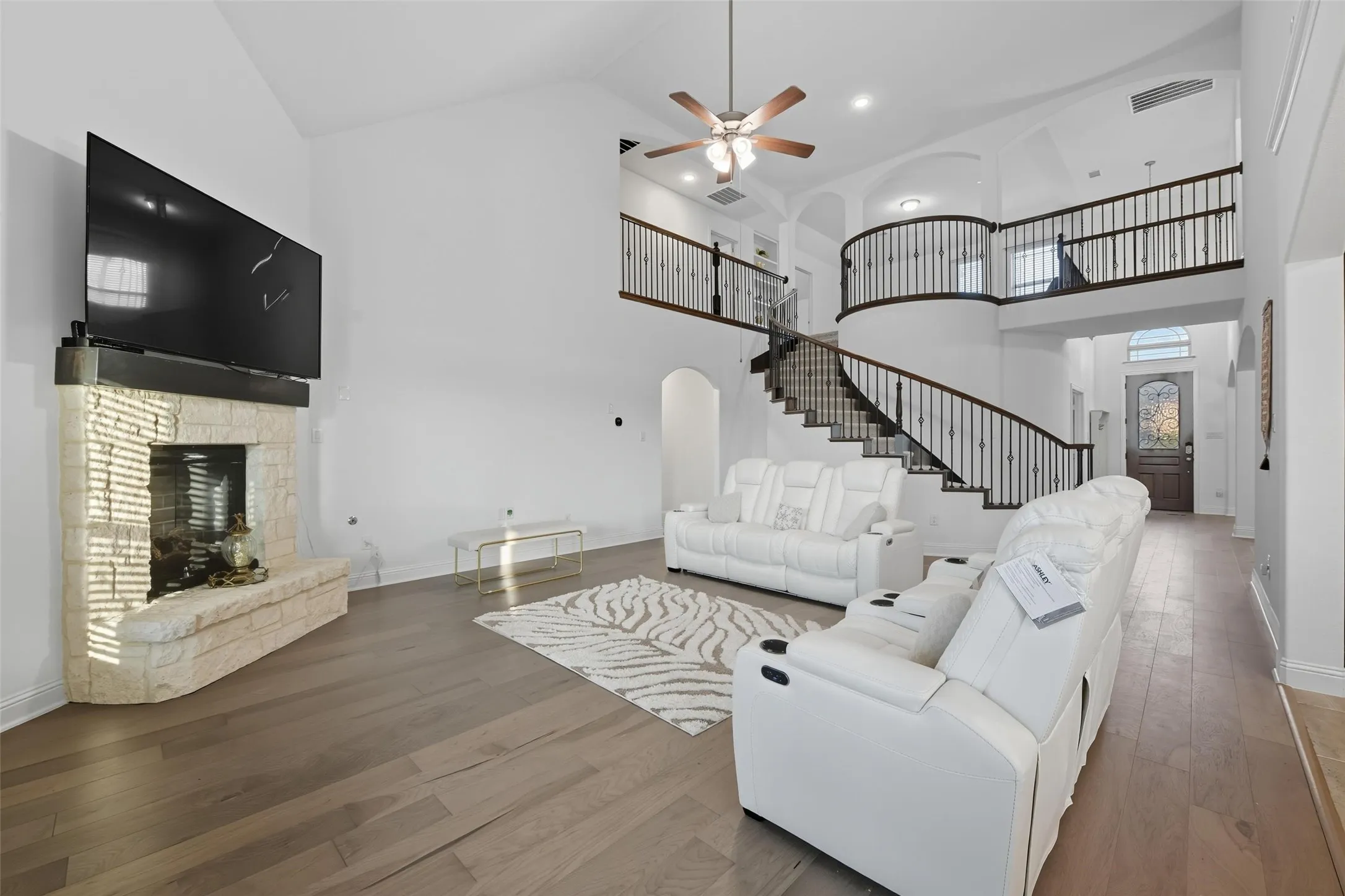 Spacious living area featuring a stone-clad fireplace, soaring vaulted ceilings, wood-finish flooring, and a grand curved staircase with wrought iron balusters