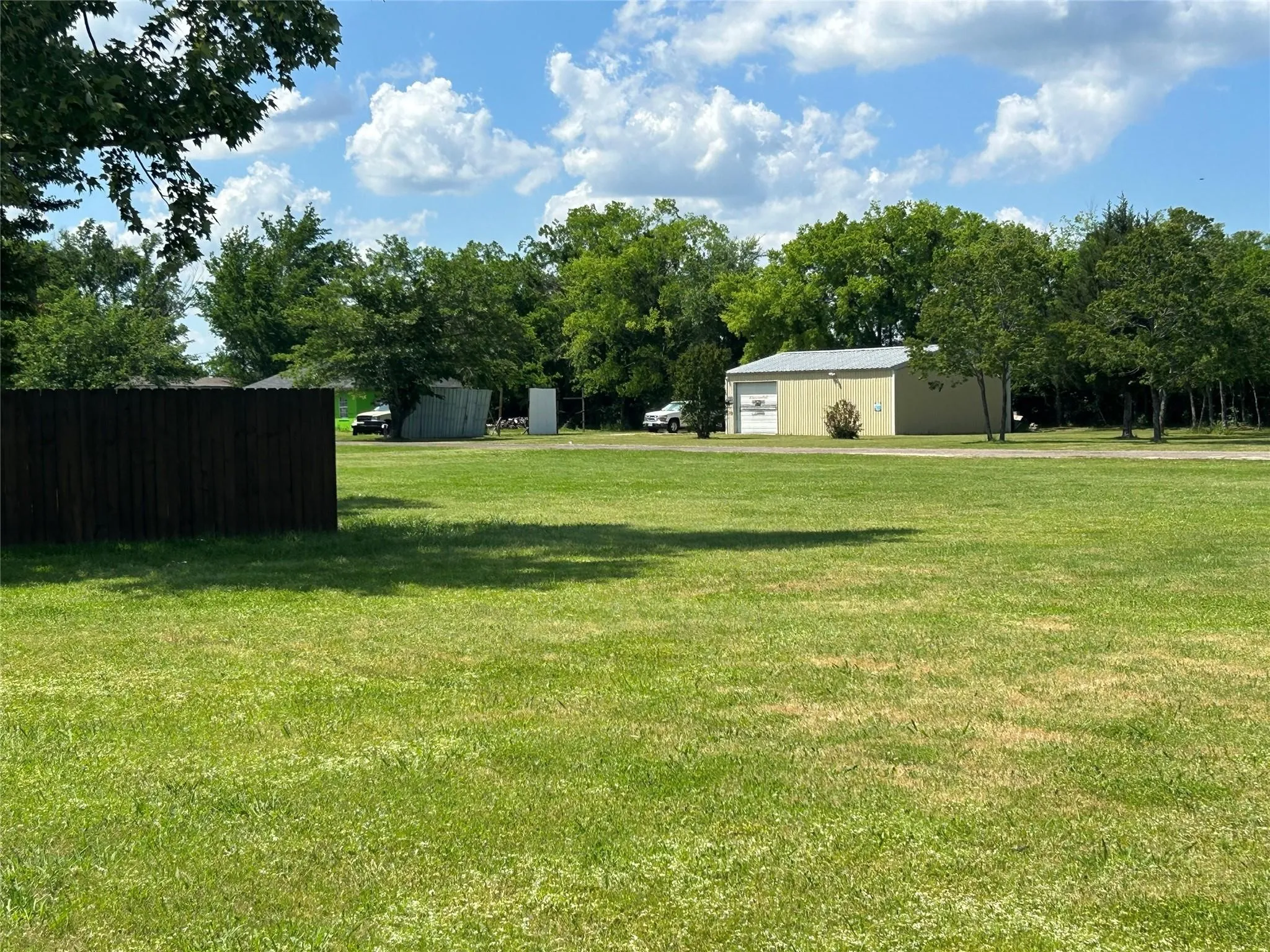 Expansive grassy yard with a detached light-colored building featuring a large garage door, surrounded by mature trees and a dark-stained wooden fence