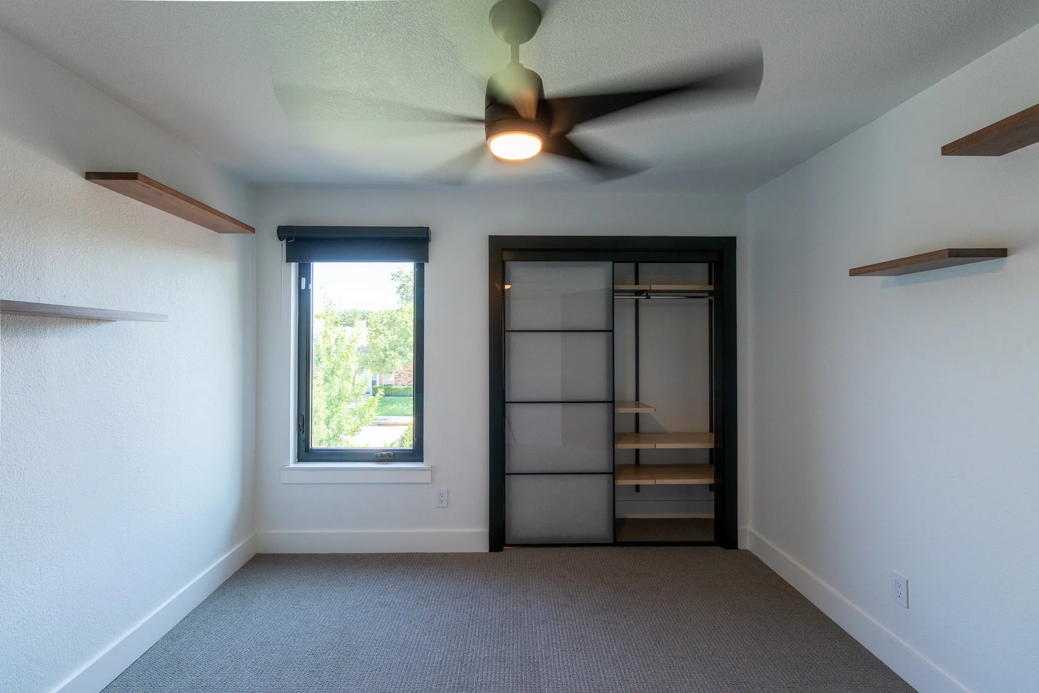 Bedroom featuring a ceiling fan with integrated lighting, a window with a dark roller shade, built-in floating shelves, and a closet with sliding panel doors and interior shelving