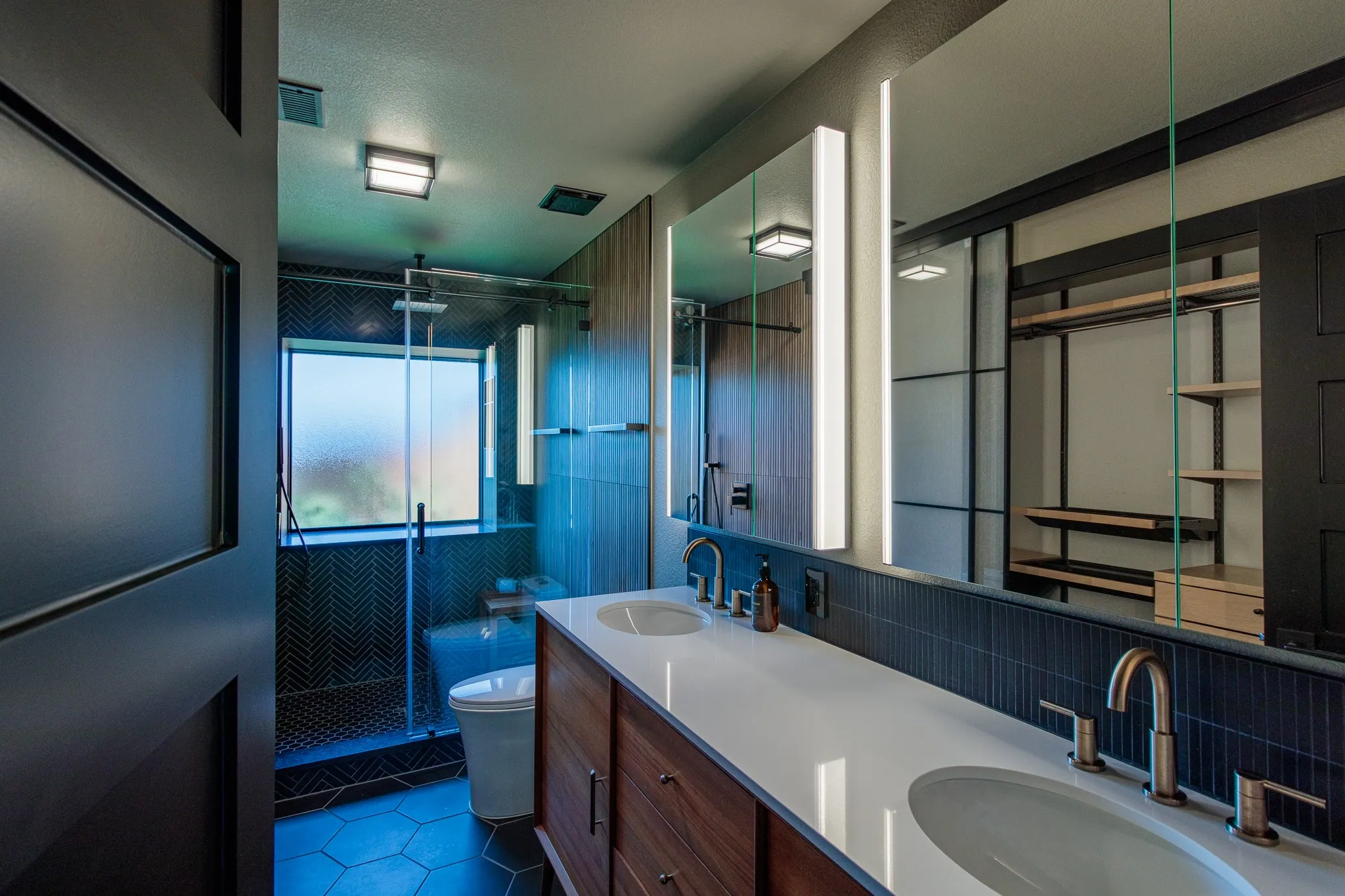 Contemporary bathroom featuring a dual vanity with white countertops and undermount sinks, wood-finish cabinetry, and a full-width mirror with integrated lighting