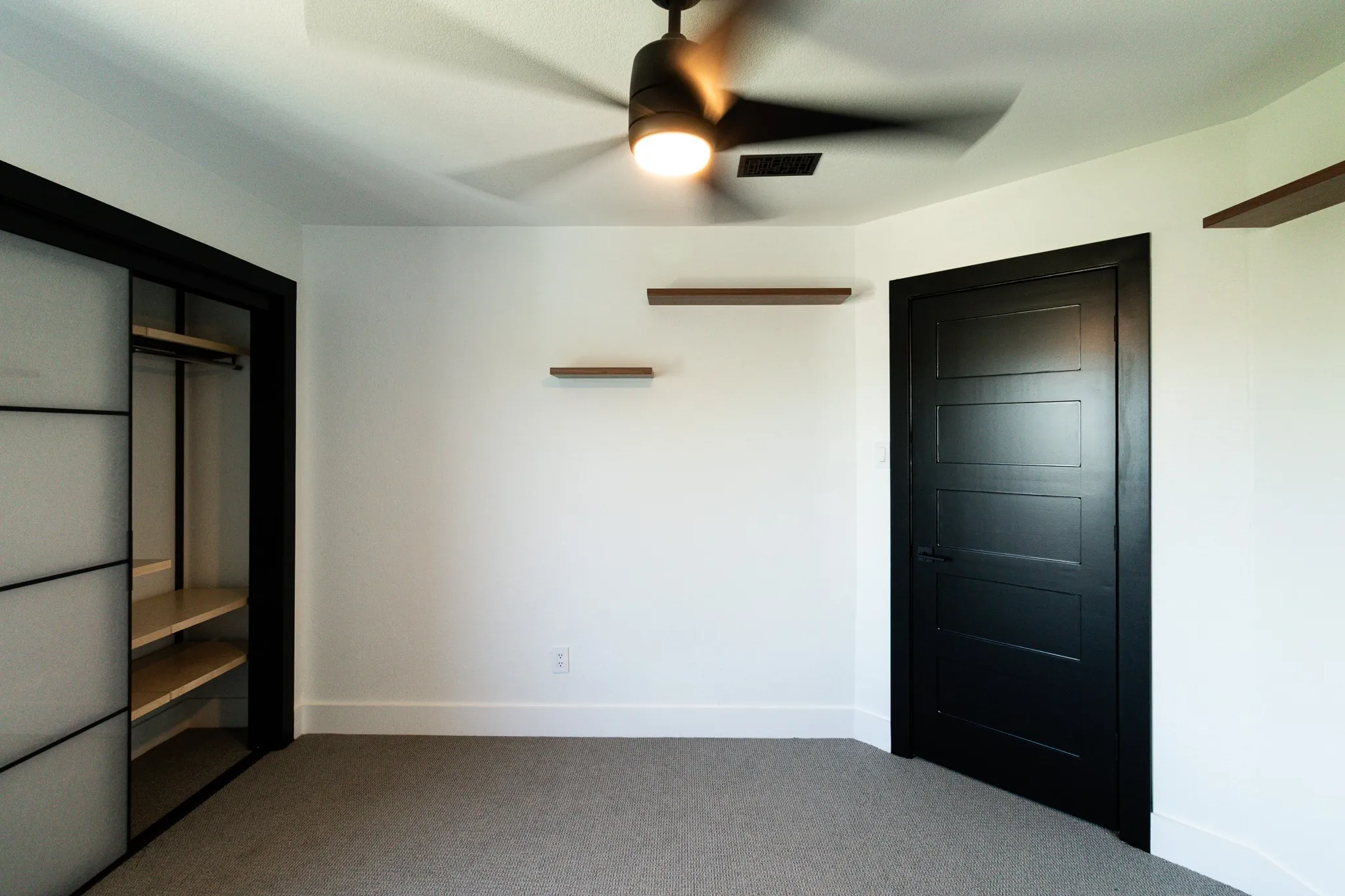 Bright interior space featuring a ceiling fan with integrated lighting, wood-finish floating shelves, and a dark paneled door with matching trim