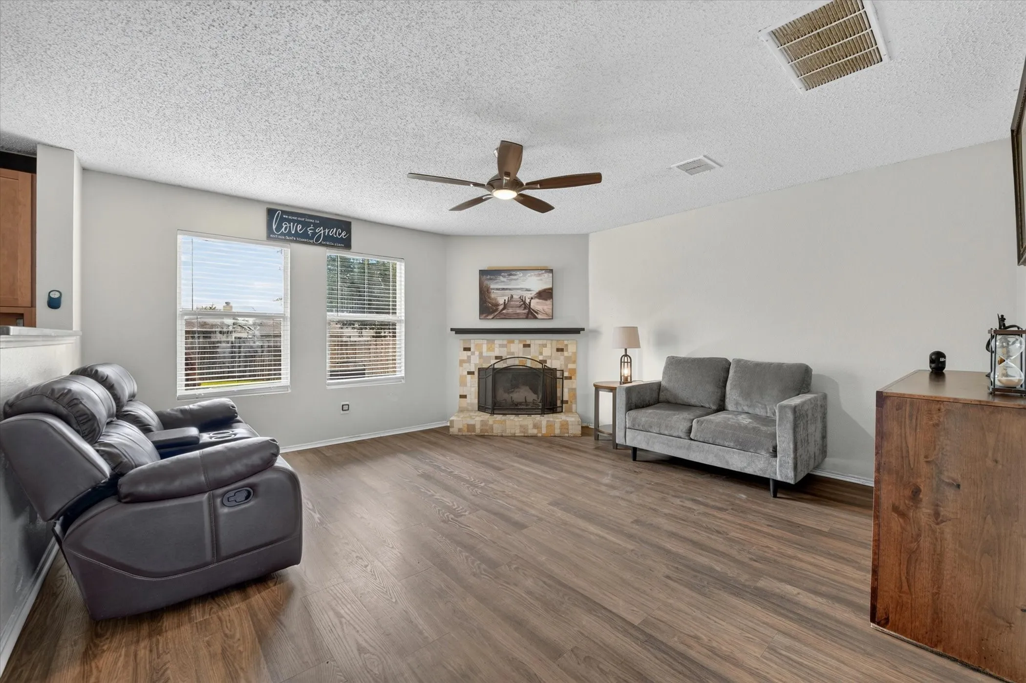 Spacious room featuring wood-finish flooring, a corner fireplace with a light-toned brick surround and black mantel, and multiple windows with blinds