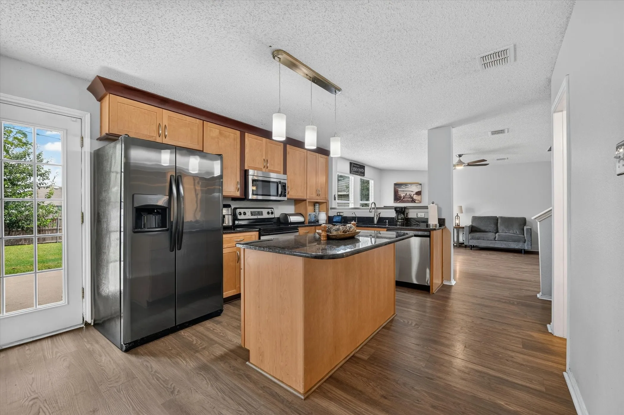Kitchen featuring wood-finish flooring, light wood cabinetry, stainless steel appliances, a center island with a dark countertop, and a glass-paneled door leading to the backyard