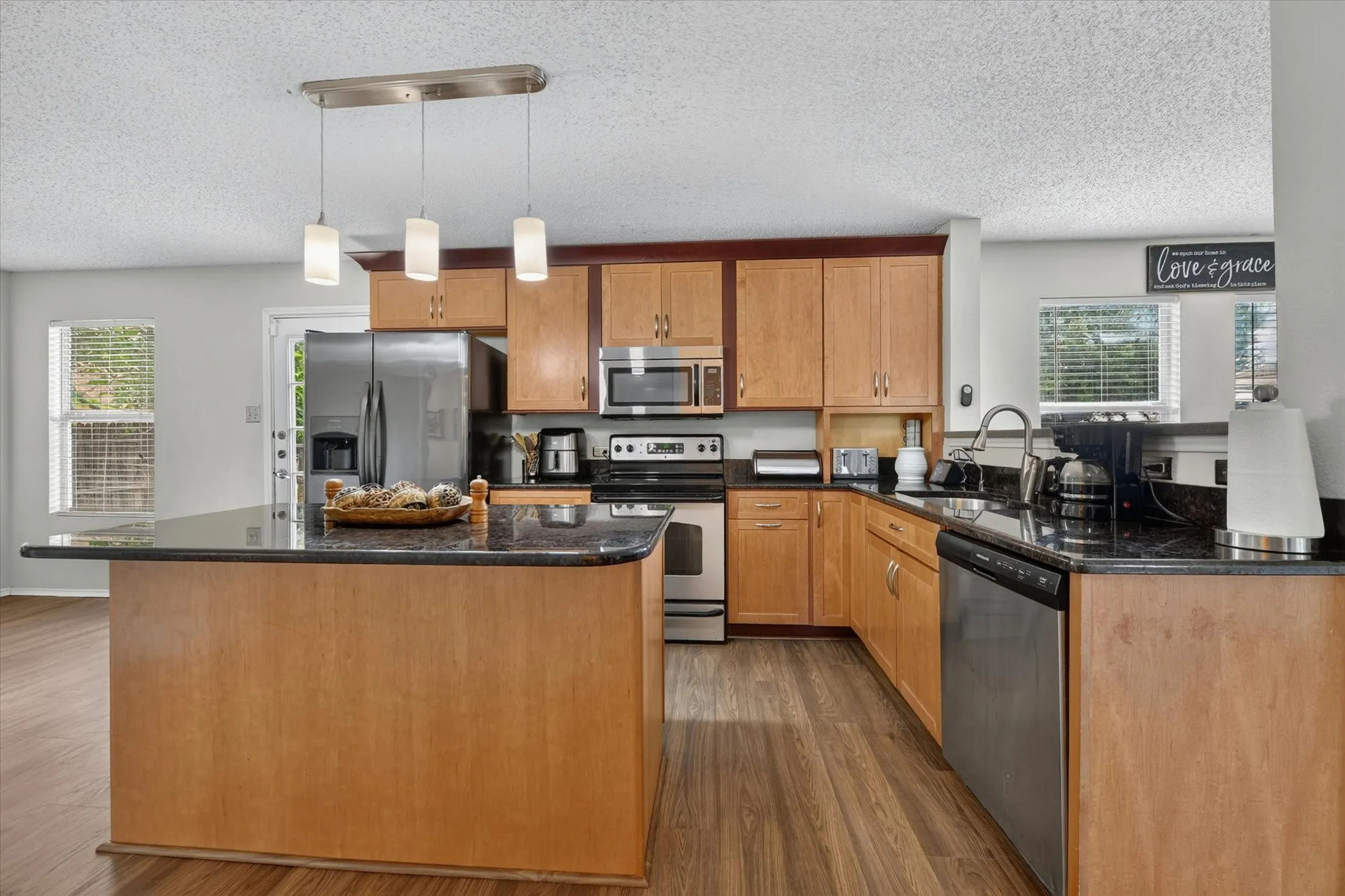 Open-concept kitchen featuring an island, wood-finish flooring, light wood cabinetry, dark countertops, and stainless steel appliances