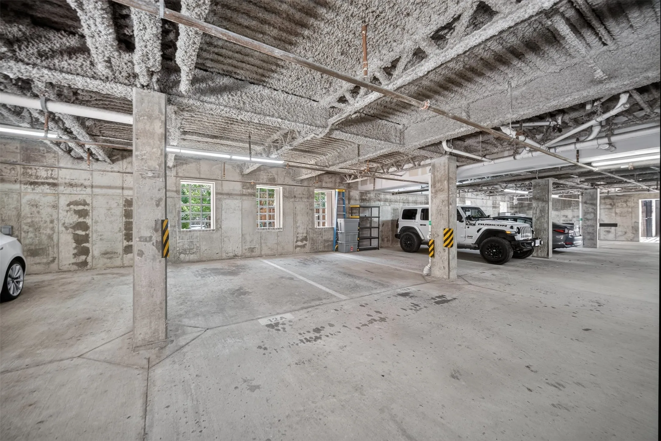Spacious concrete parking garage featuring exposed overhead ductwork, multiple support columns, perimeter windows, and overhead strip lighting