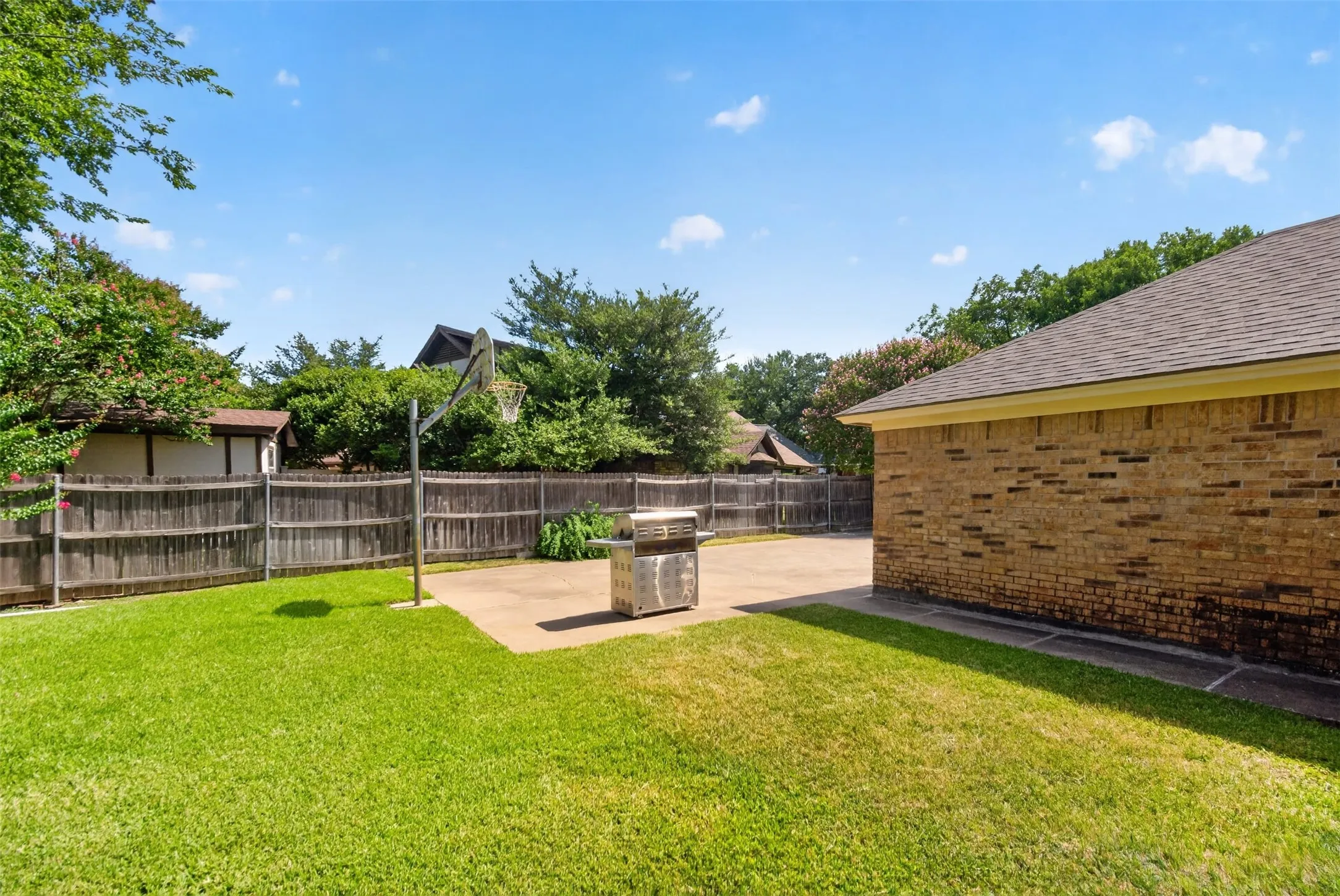 Expansive backyard featuring a concrete patio, a basketball hoop, and a brick exterior wall