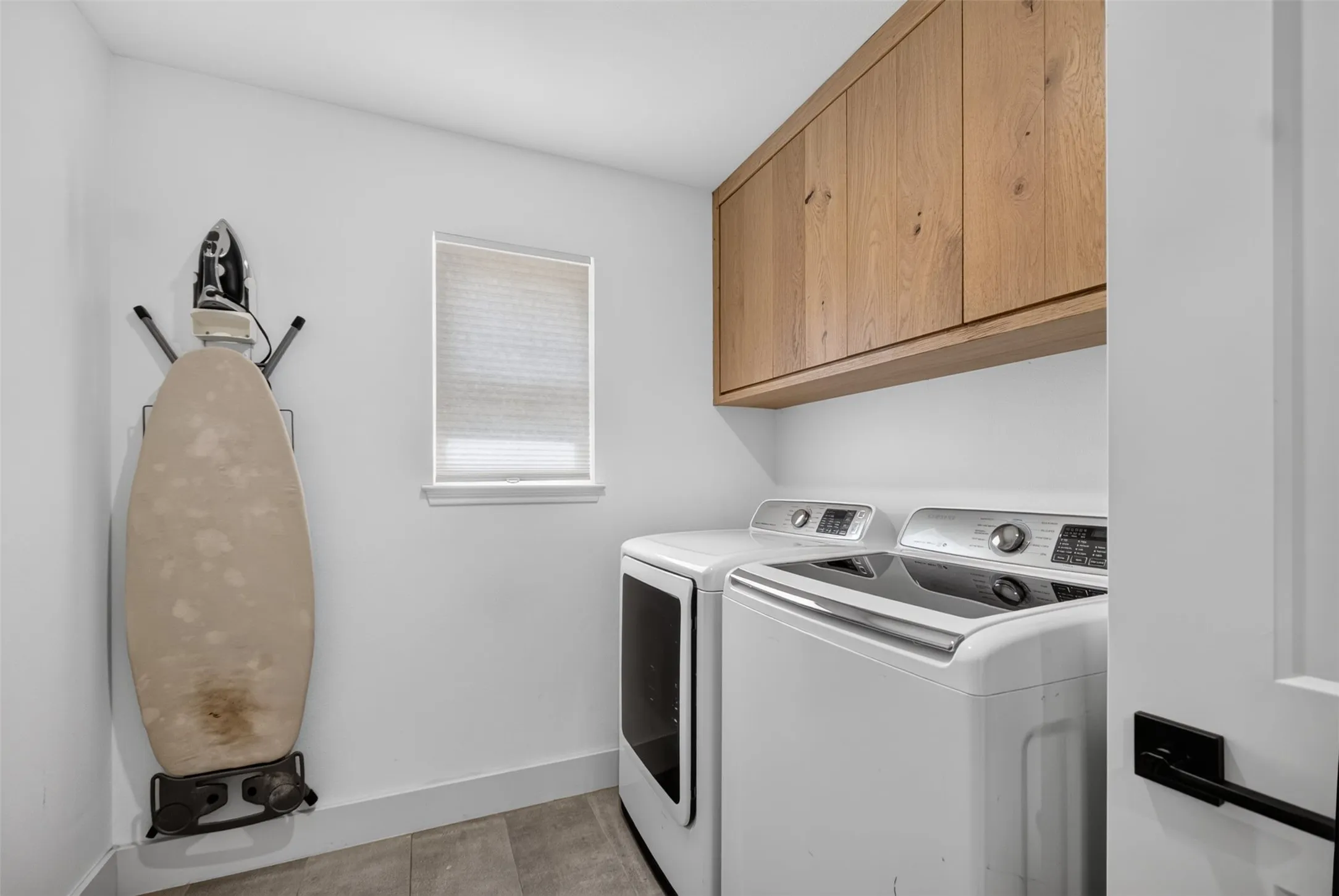 Dedicated laundry area featuring wood-finish upper cabinetry, a window with blinds, and tile flooring