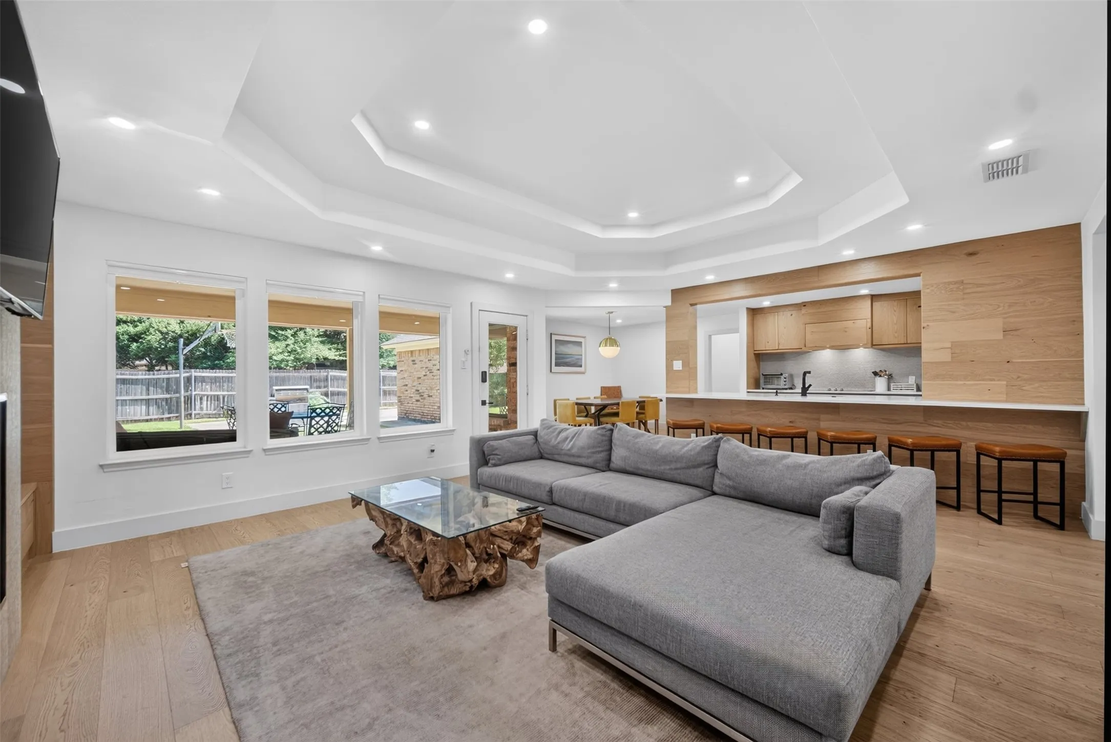 Open-concept living area with tray ceiling, recessed lighting, wood-finish flooring, and a kitchen pass-through featuring light wood cabinetry