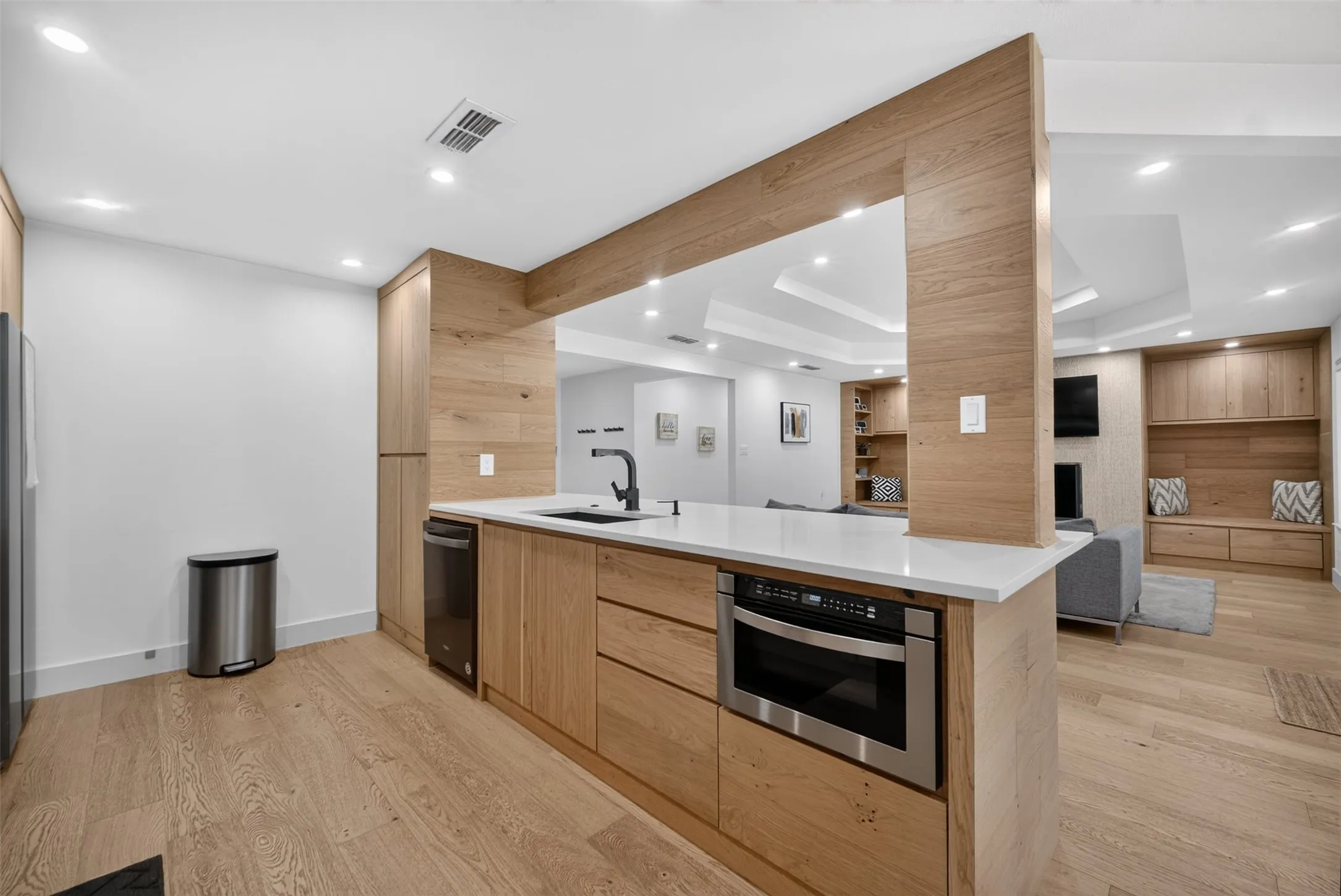Modern kitchen featuring light wood-finish cabinetry, white countertops, a built-in stainless steel microwave, a black sink with a matte black faucet, and wood-finish flooring