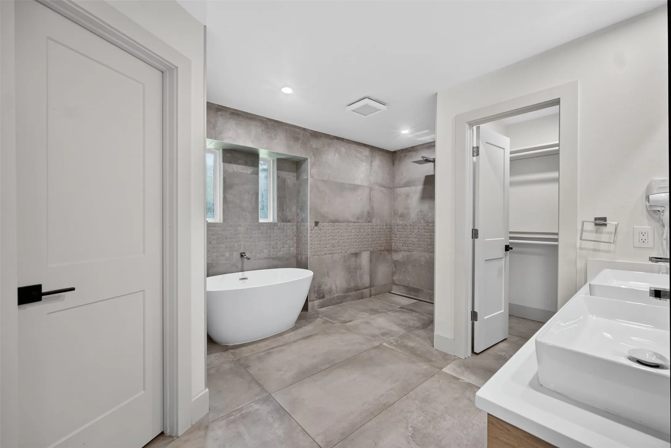 Full bathroom featuring a freestanding soaking tub, a walk-in shower with large format tile, dual vessel sinks on a vanity with a light wood-finish base, and a walk-in closet with built-in shelving