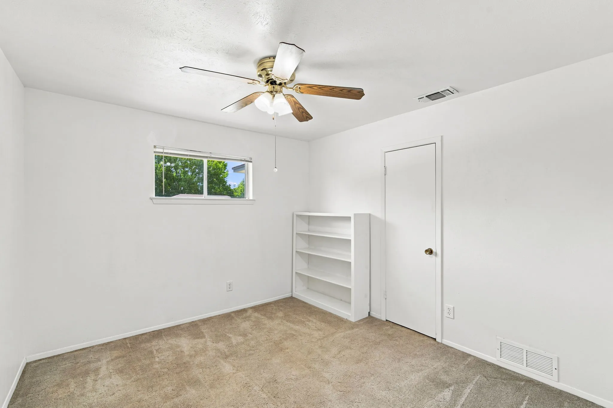 Carpeted room featuring a ceiling fan, a window with exterior views, a built-in white shelving unit, and a white interior door